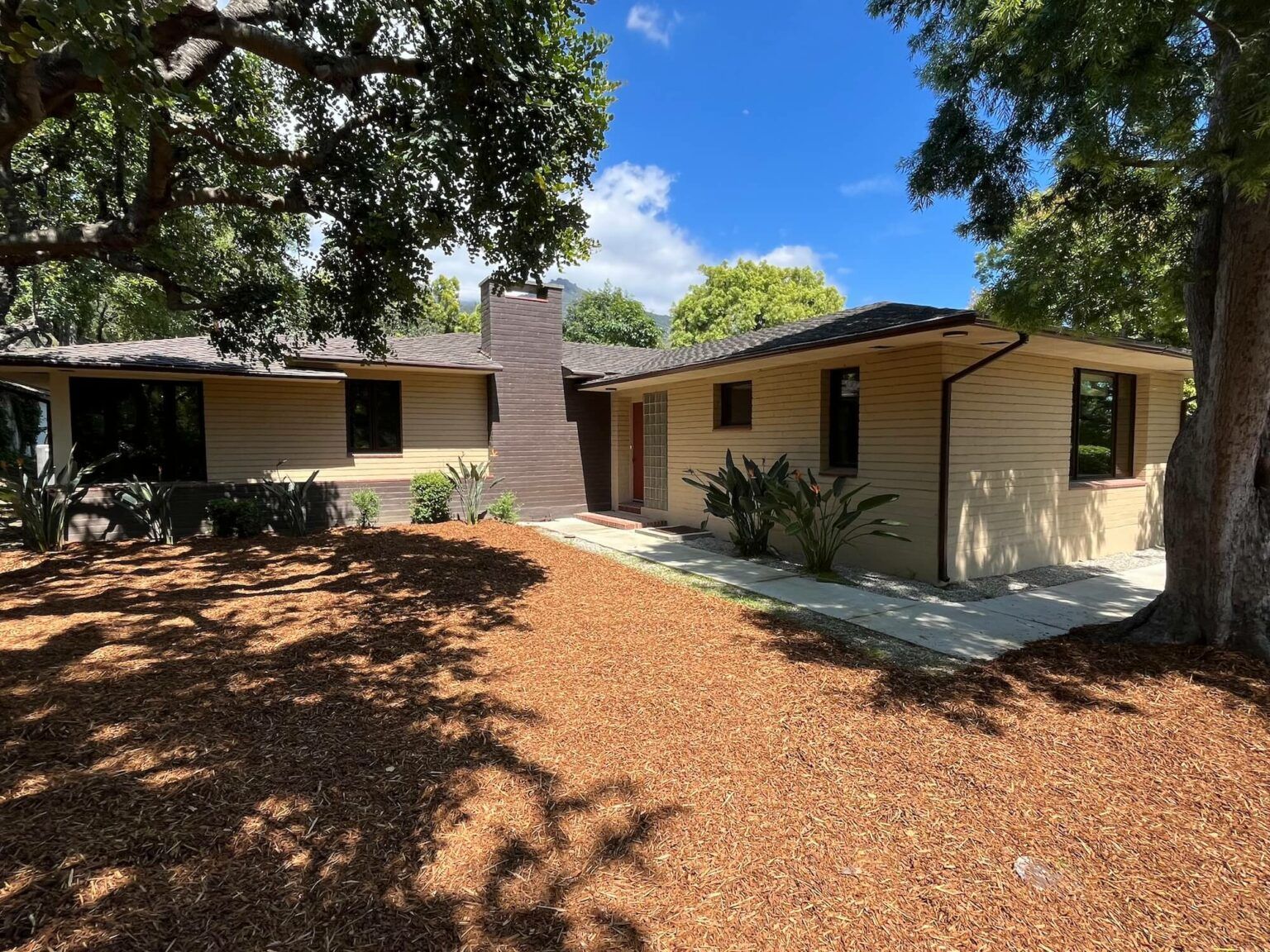 Beige ranch-style house with dark-trimmed windows, brown roof, and brick chimney, under a tree canopy.