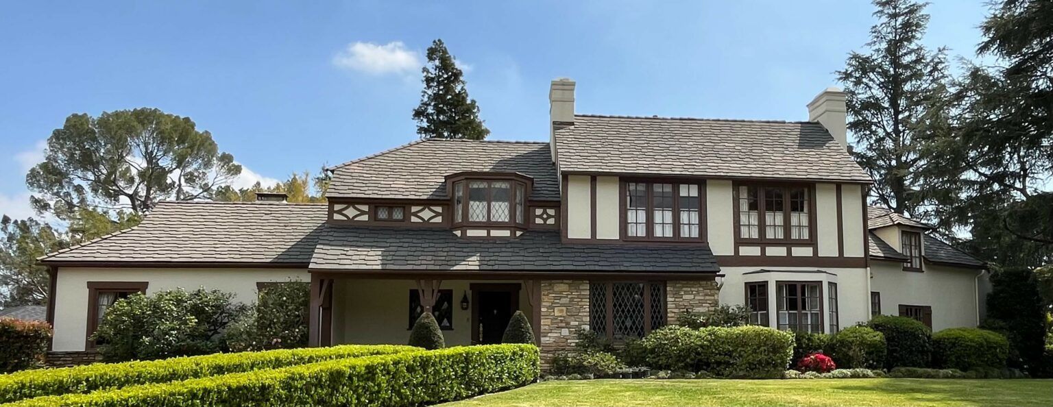 Large Tudor-style house with stone and timber accents, surrounded by a green lawn and manicured hedges, under a blue sky.