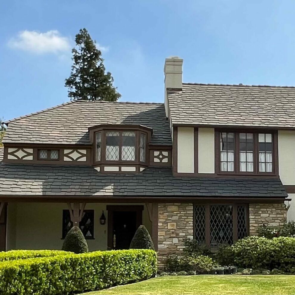 Tudor-style house with stone facade, dark-brown trim, and a shingled roof, set against a blue sky.