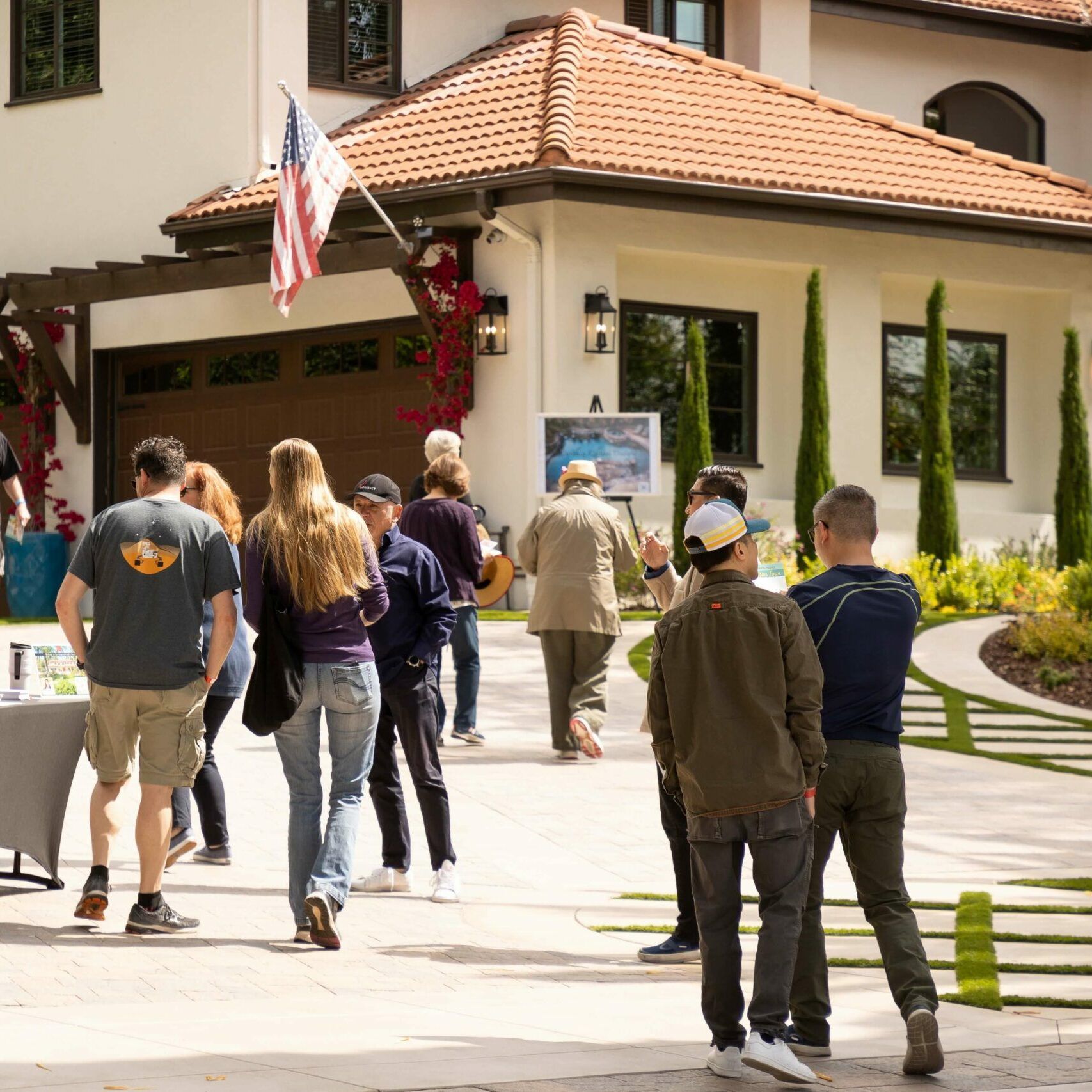 People gather outside a house with a flag, some looking at displayed art.