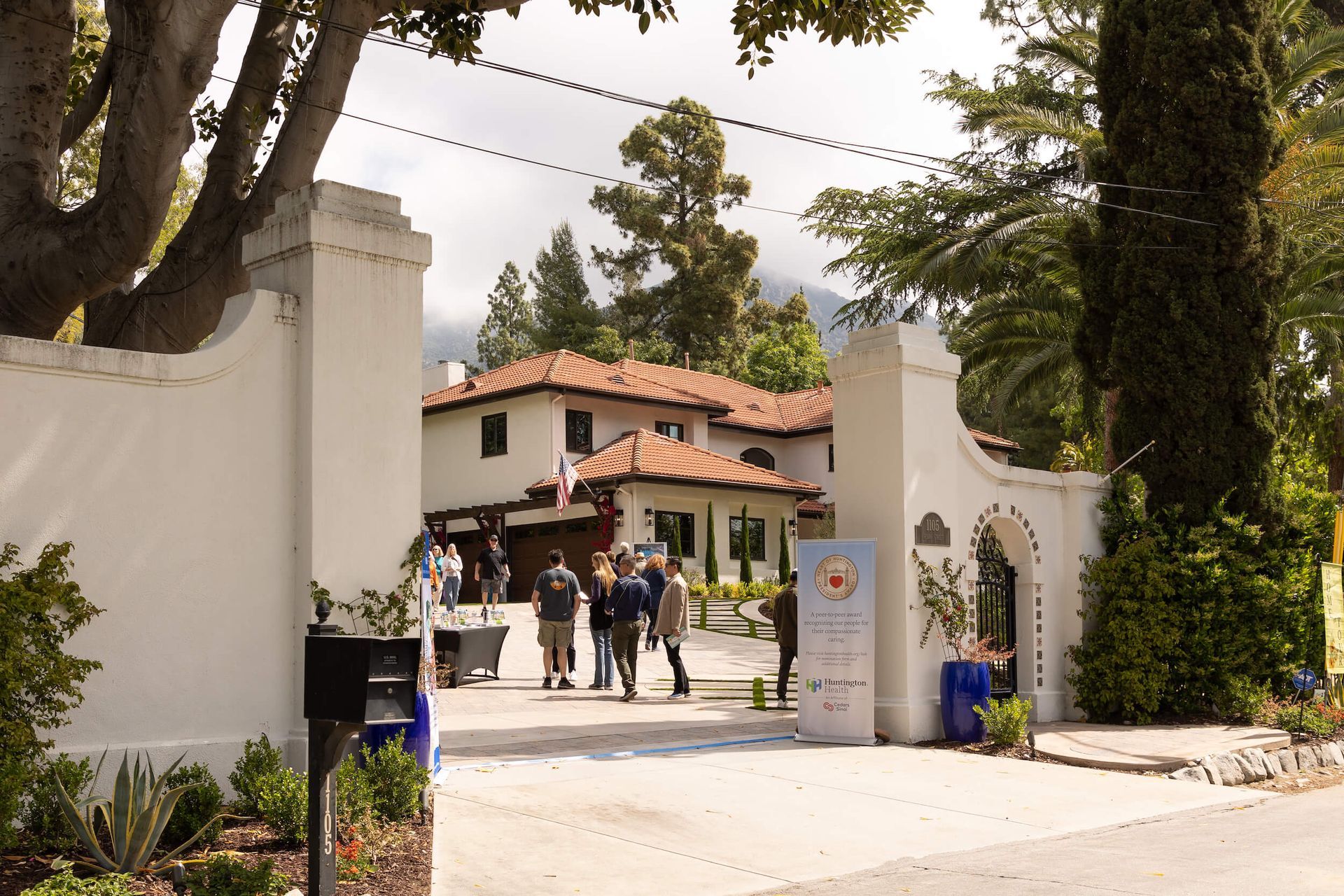 White-walled entrance to a Spanish-style building; people gathered, trees frame the view, cloudy sky.