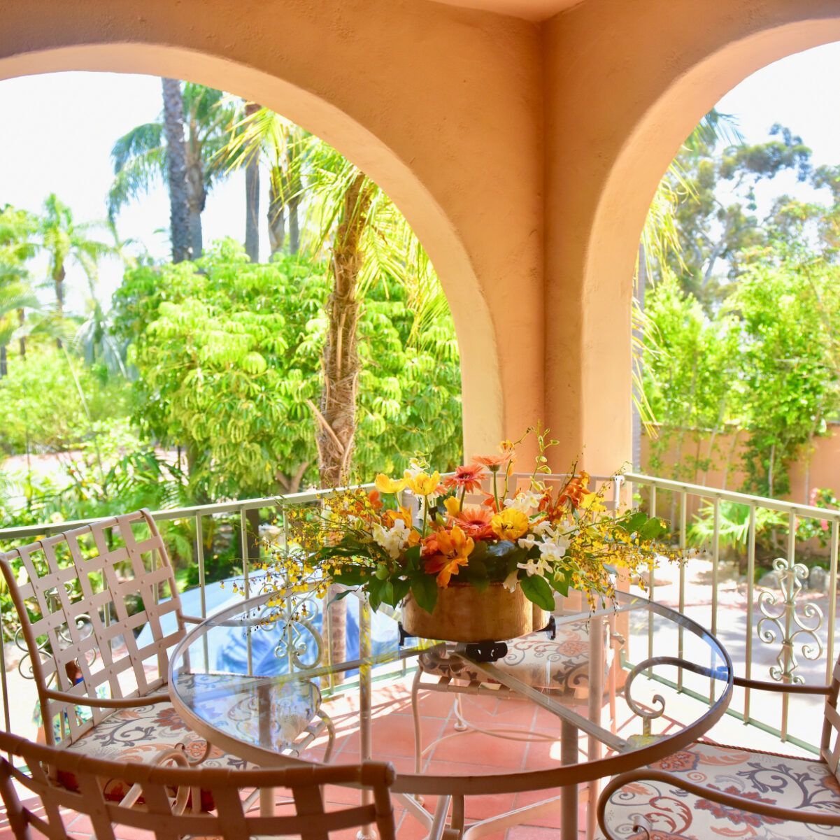 Outdoor patio with glass table, chairs, and flower arrangement, overlooking lush green foliage.