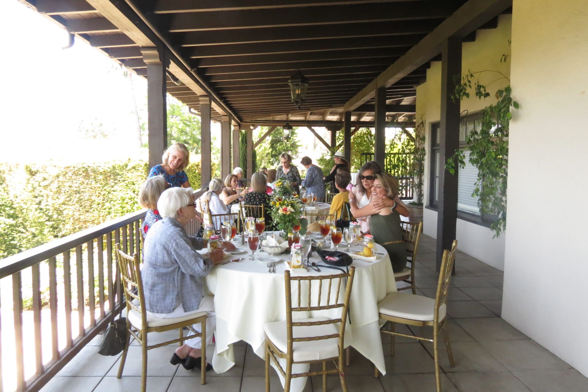People seated at round tables on a covered porch, enjoying a meal. Sunlight, greenery, and brown wood are visible.