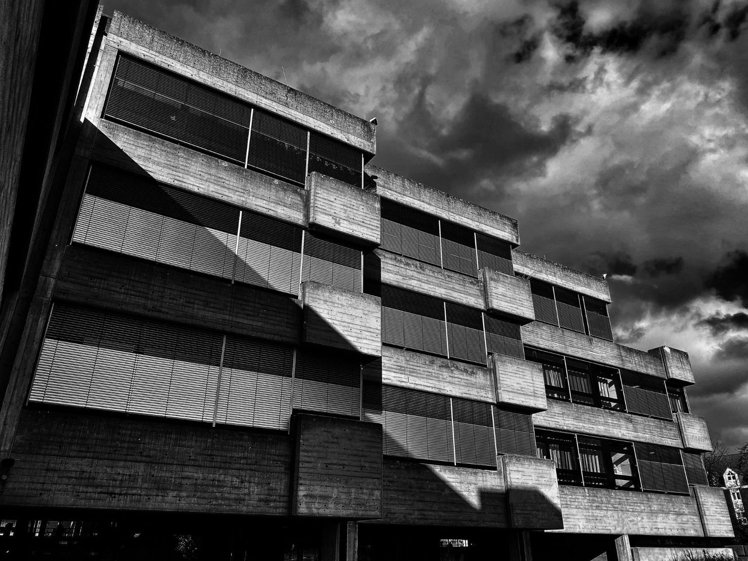 A low-angle, black-and-white shot of a tiered, modernist brick building against a dramatic, cloudy sky.