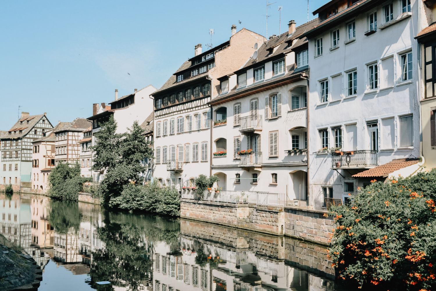 Gebäude entlang eines Kanals in Straßburg, Frankreich; helle Fassaden, Spiegelungen im Wasser, blauer Himmel.