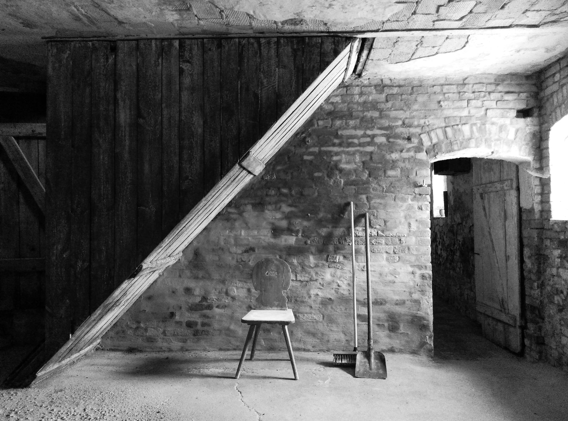 Schwarz-weißes Interieur mit Holztür, schrägem Stützbalken, Hocker, Schaufel und Ziegelwänden.