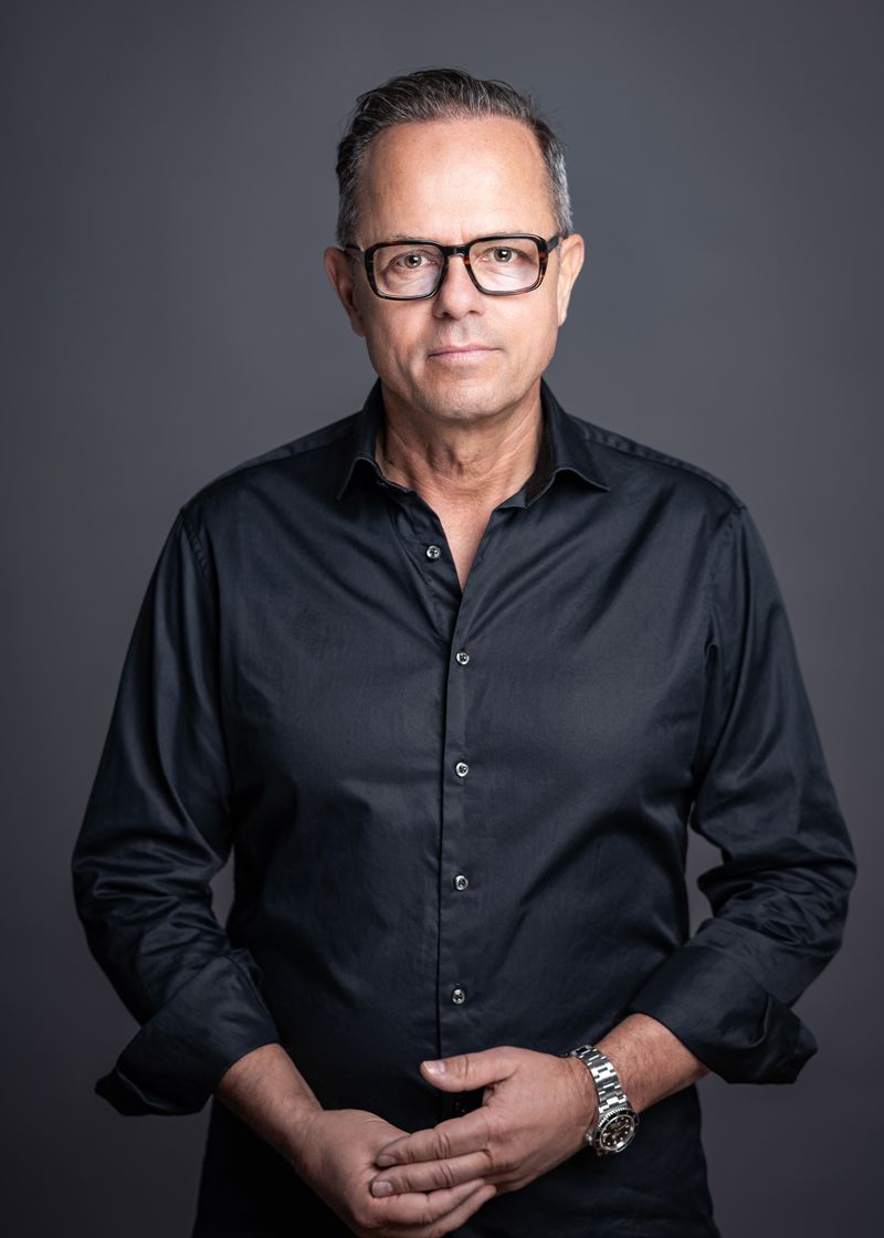 Mann mit Brille, schwarzem Hemd und verschränkten Armen vor grauem Hintergrund.