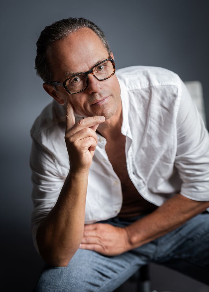 Mann mit Brille, Hand am Kinn, trägt ein weißes Hemd und Jeans, sitzt und sieht nachdenklich aus.