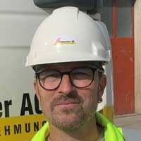 Ein Mann mit Schutzhelm und Brille steht vor einem Gebäude.