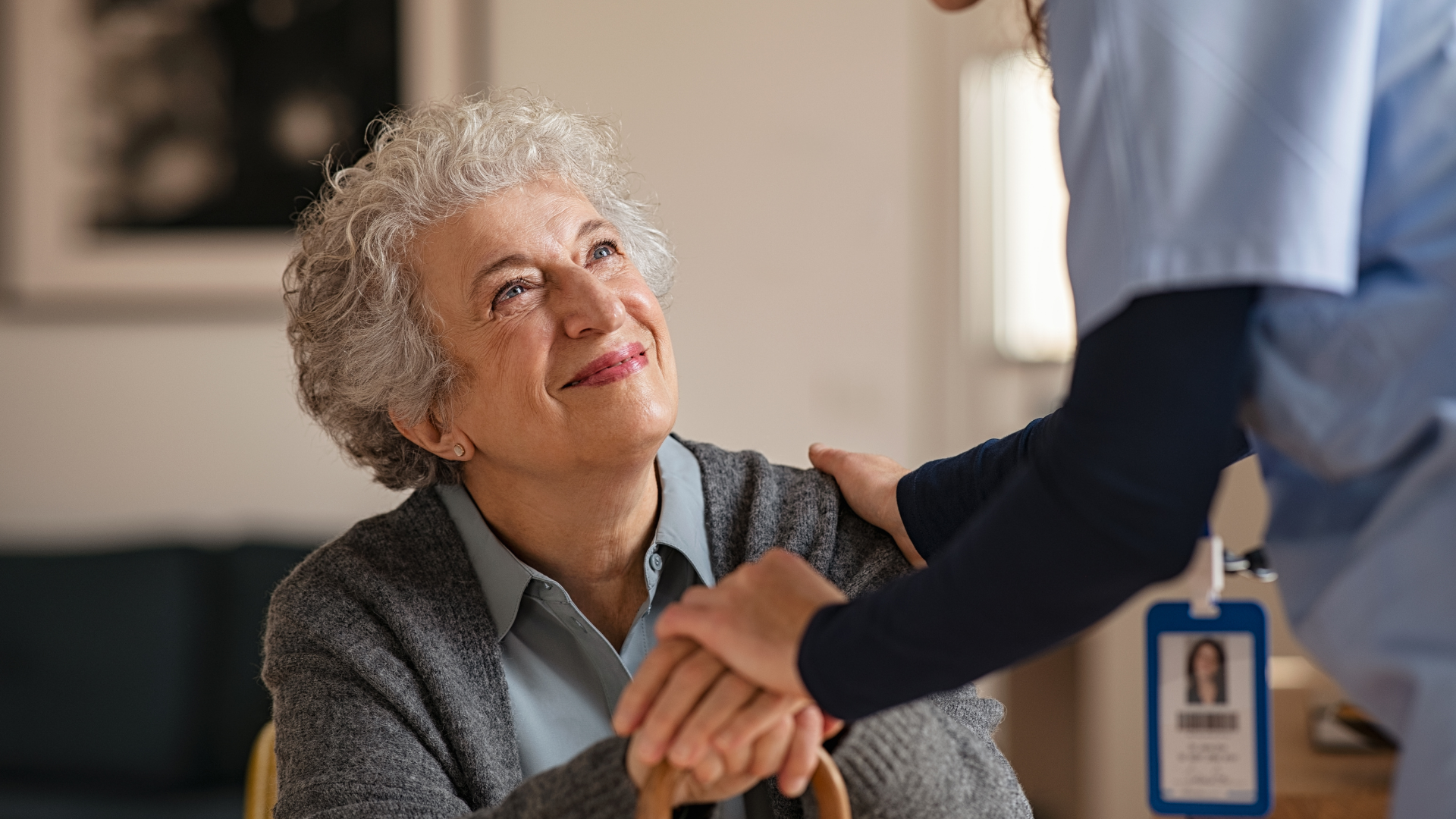 Man in blue scrubs explains medication to an older woman on a couch; they are in a home setting.