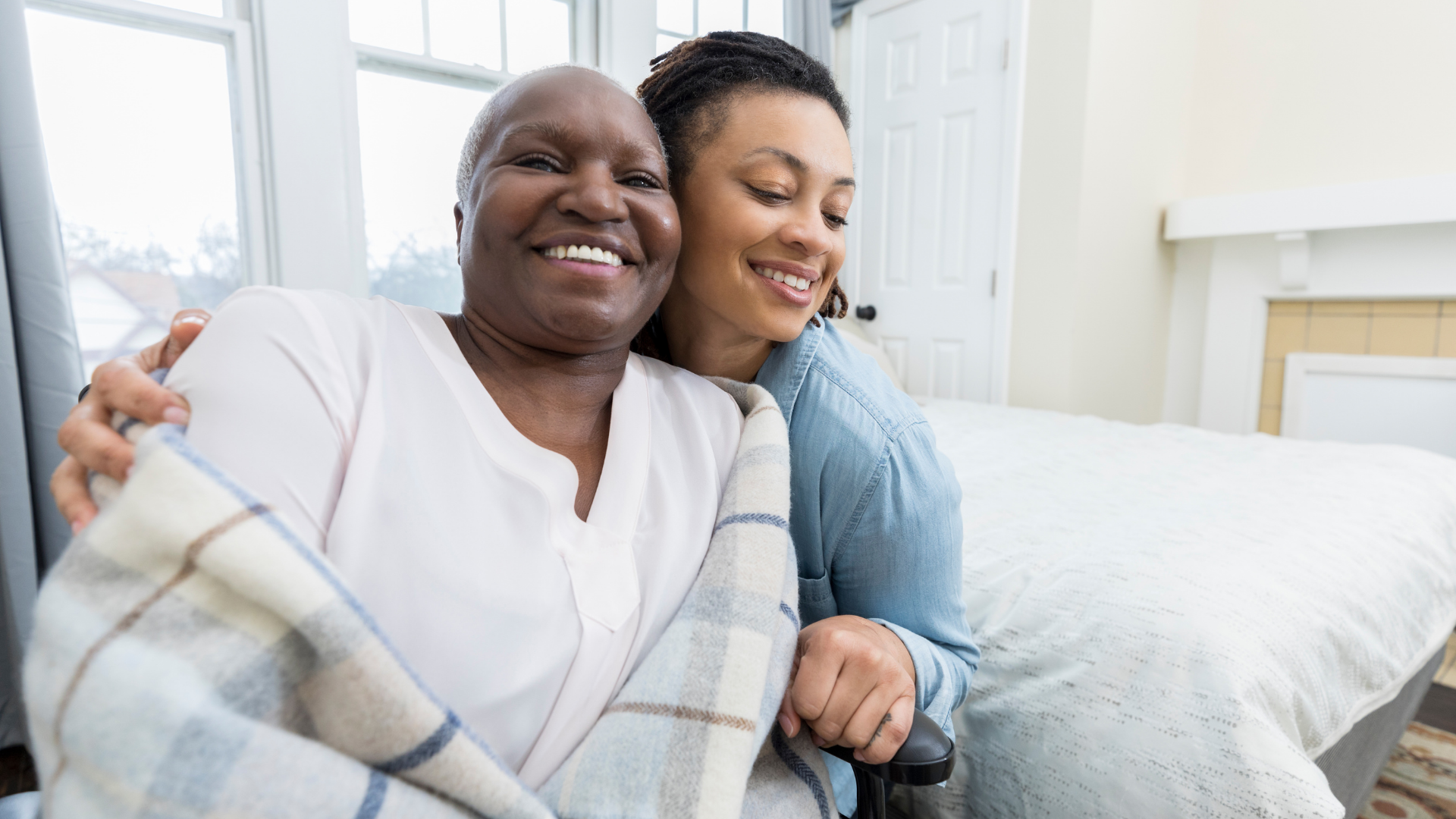 Woman in wheelchair smiles, wrapped in blanket, with another woman's arm around her in a room, near a bed.
