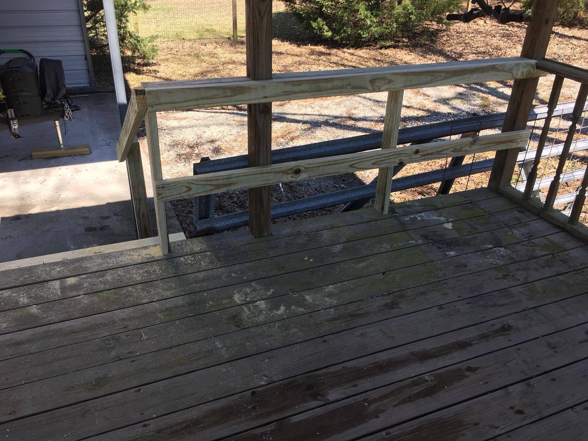 A view of a weathered wooden porch deck with a simple, unfinished wooden handrail and steps leading to the ground.
