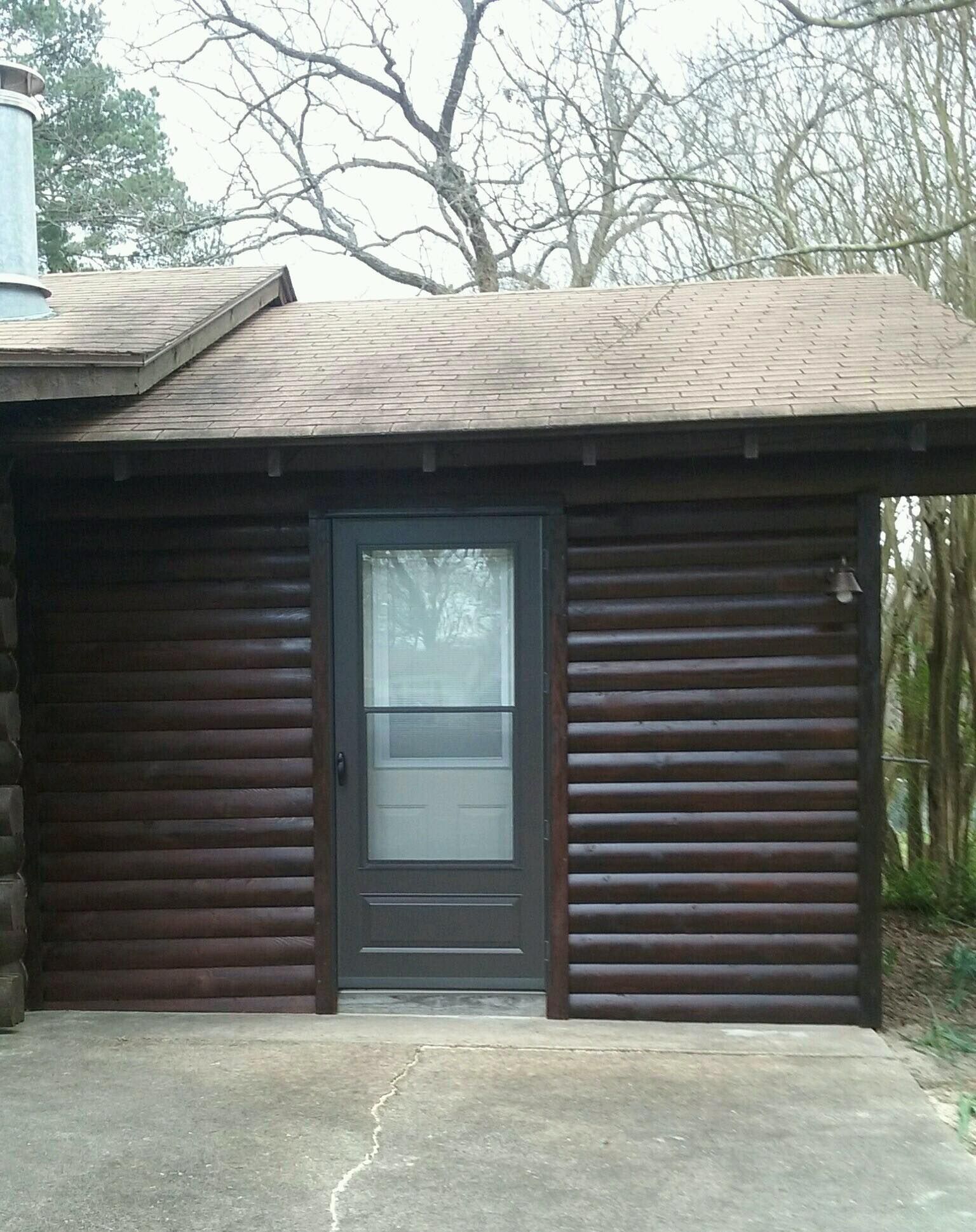 A dark wood-paneled cabin exterior with a central glass screen door under an overhanging roof, set above a concrete patio.