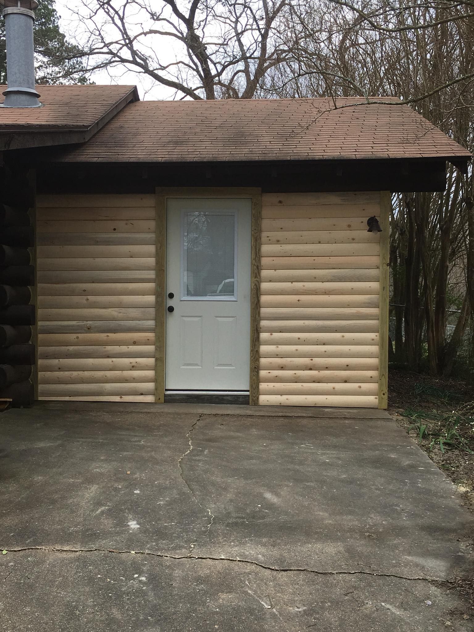 A white door centered on the exterior wall of a cabin with unfinished light wood siding and a brown shingled roof.
