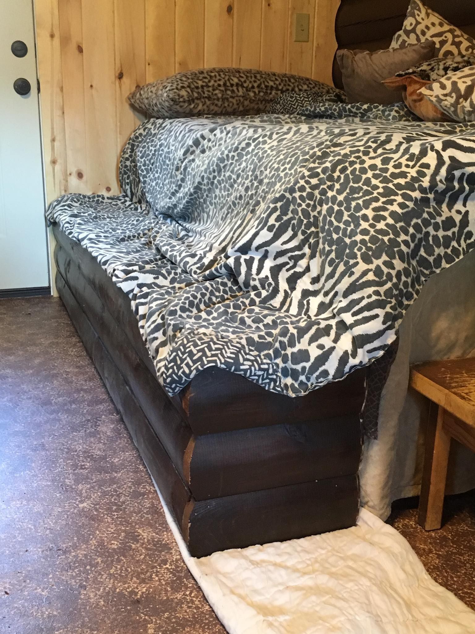 A bed with a dark wood base and patterned gray and white bedding sits in a room with light wood paneled walls.