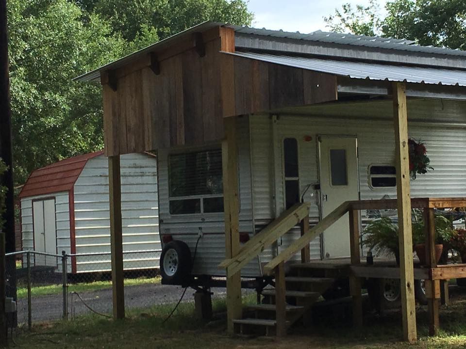 A travel trailer with a wooden extension roof and a wooden deck, situated next to a small white storage shed with a red roof.