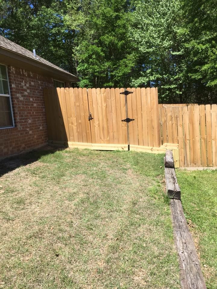 A side yard with a brick house wall, a new wooden fence with a gate, and a weathered wooden landscape timber on the grass.