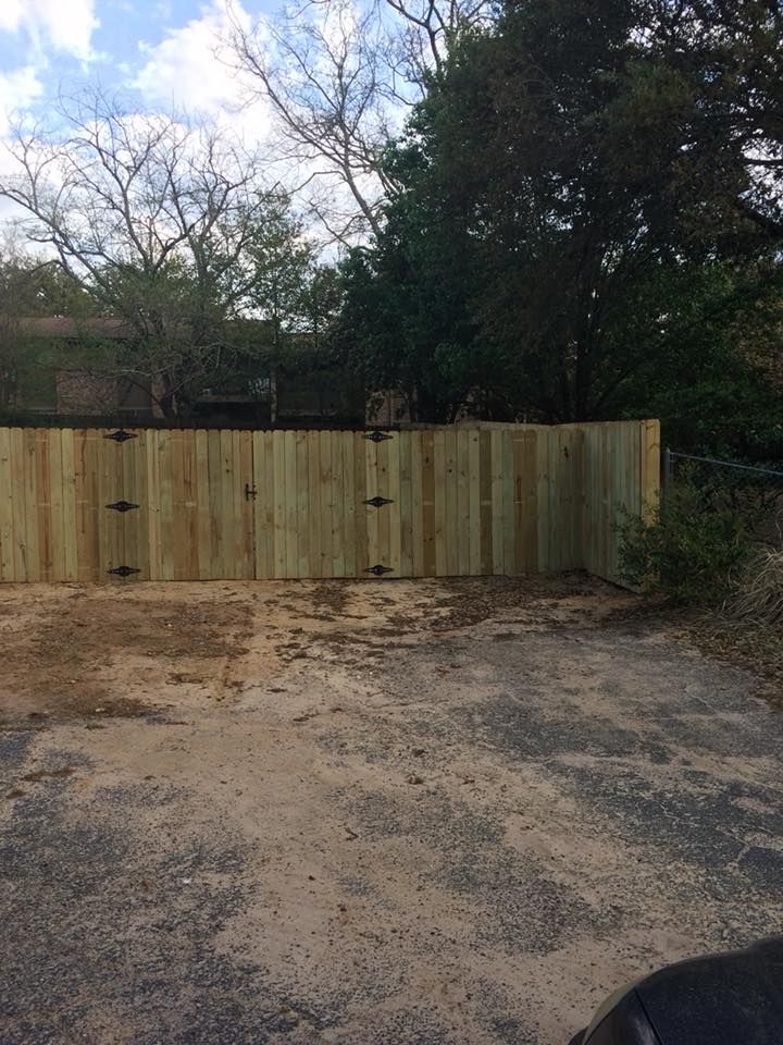 A newly installed light-wood privacy fence with double gates sits in a gravel lot with trees in the background.