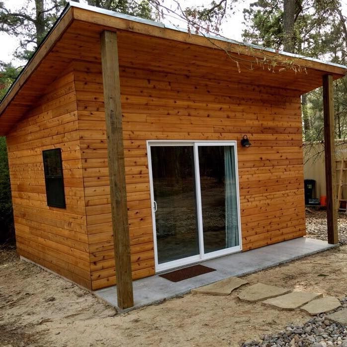 A small, modern wooden cabin with a single-slope roof, sliding glass doors, and a small concrete porch in a wooded area.