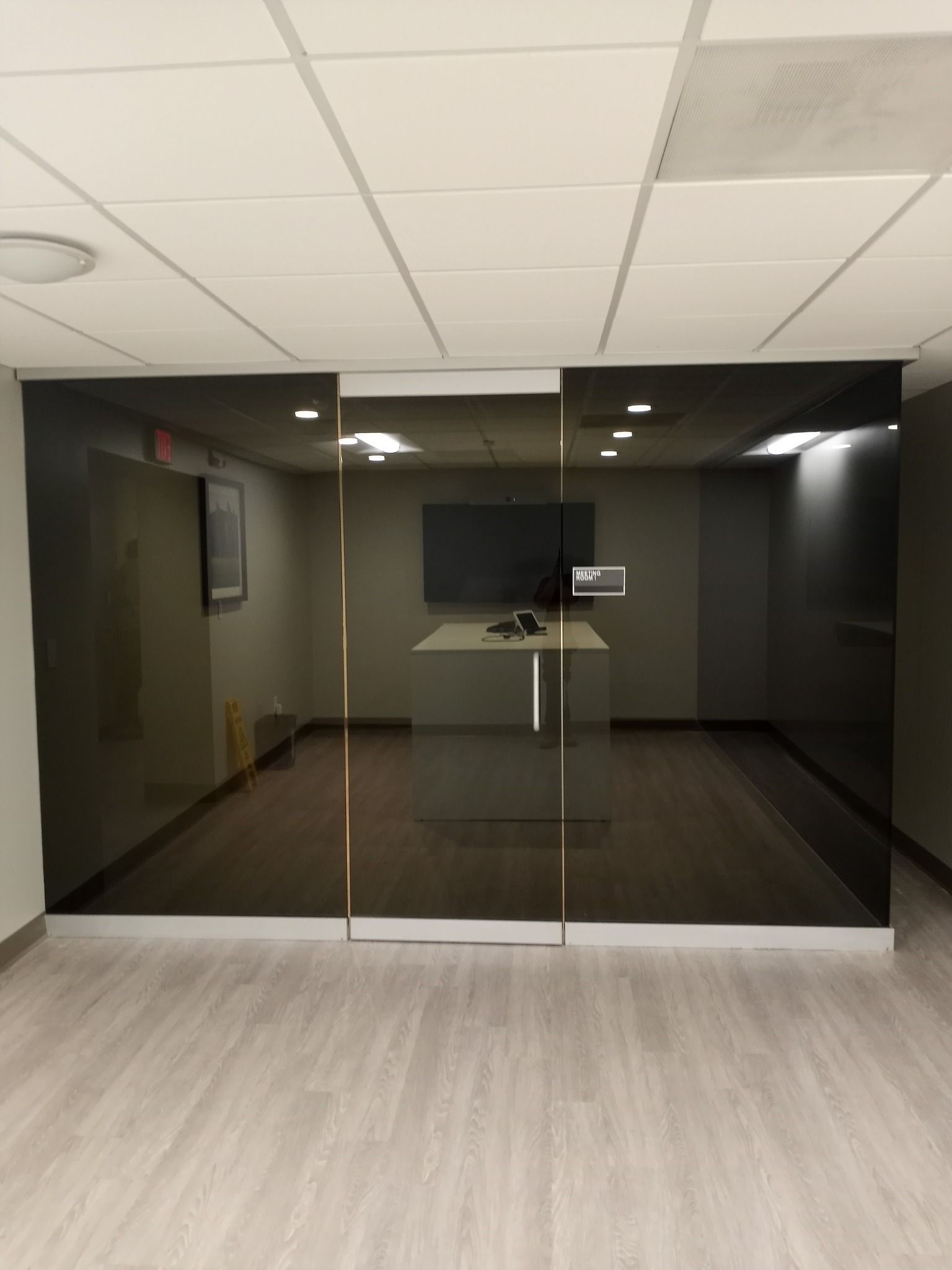 Glass-walled office space with a rectangular table, screen, and dark gray walls. Light wooden floor and white ceiling.