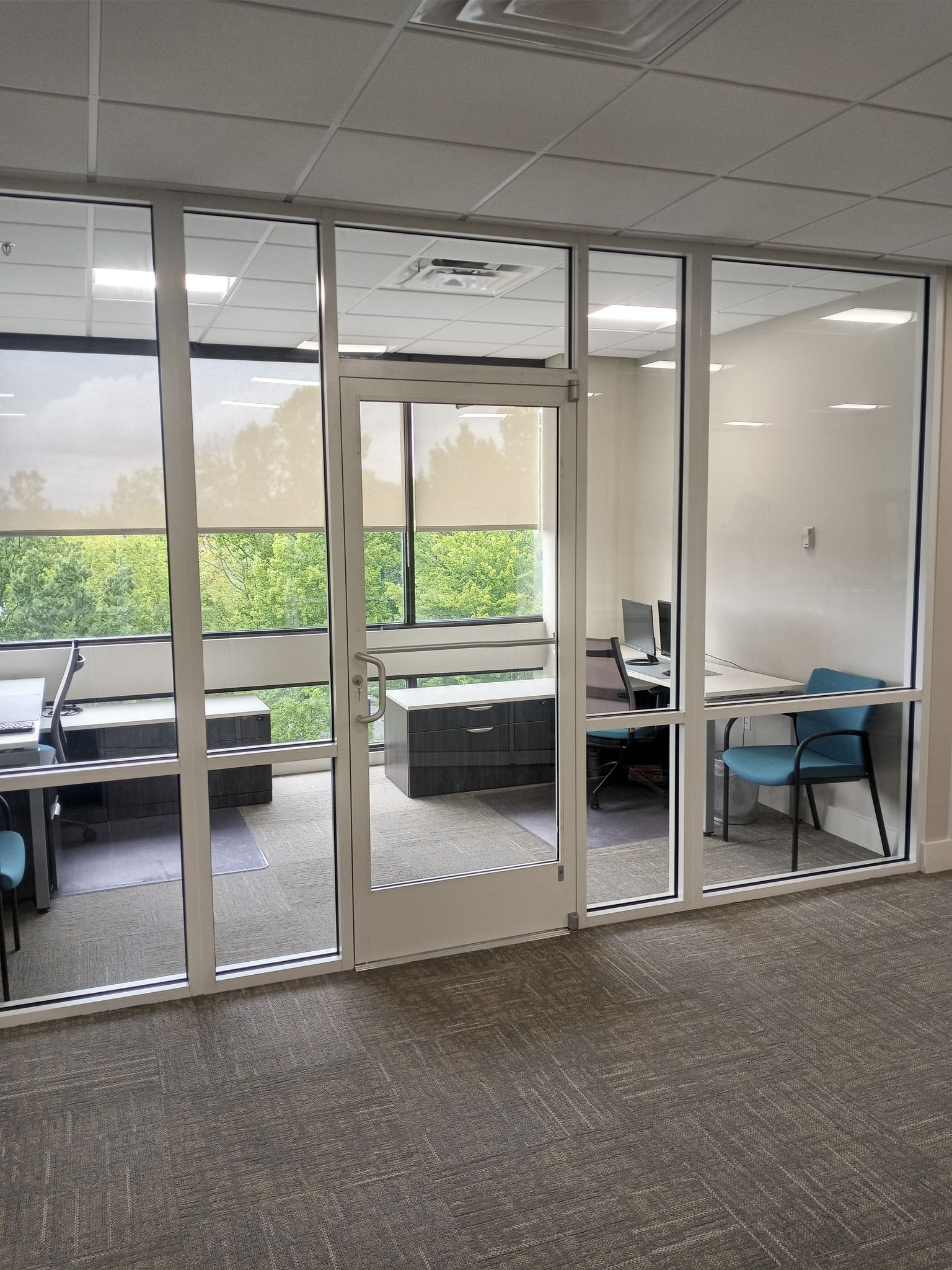 Office interior with glass walls, door, and desks. Carpeted floor, white ceiling, and natural light from outside.