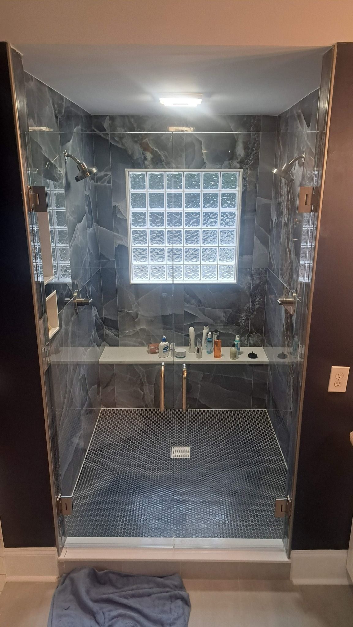 Glass-enclosed shower with patterned dark gray walls, glass block window, and various toiletries on shelf.