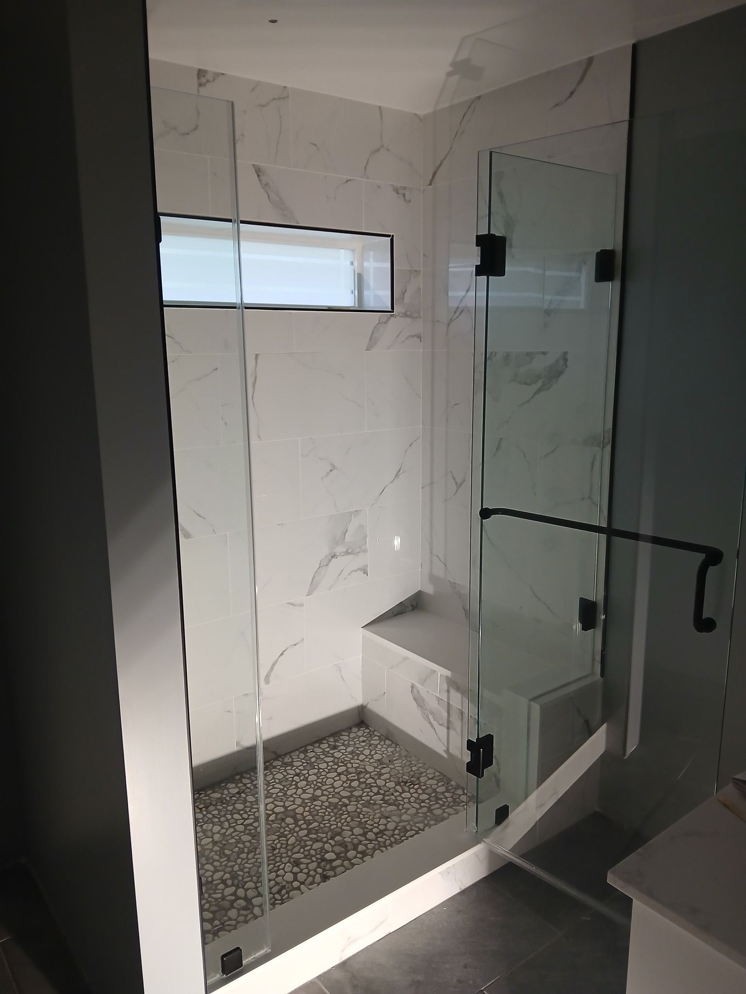 Shower stall with glass doors, marble-look tile, and pebble floor. A white bench sits inside.