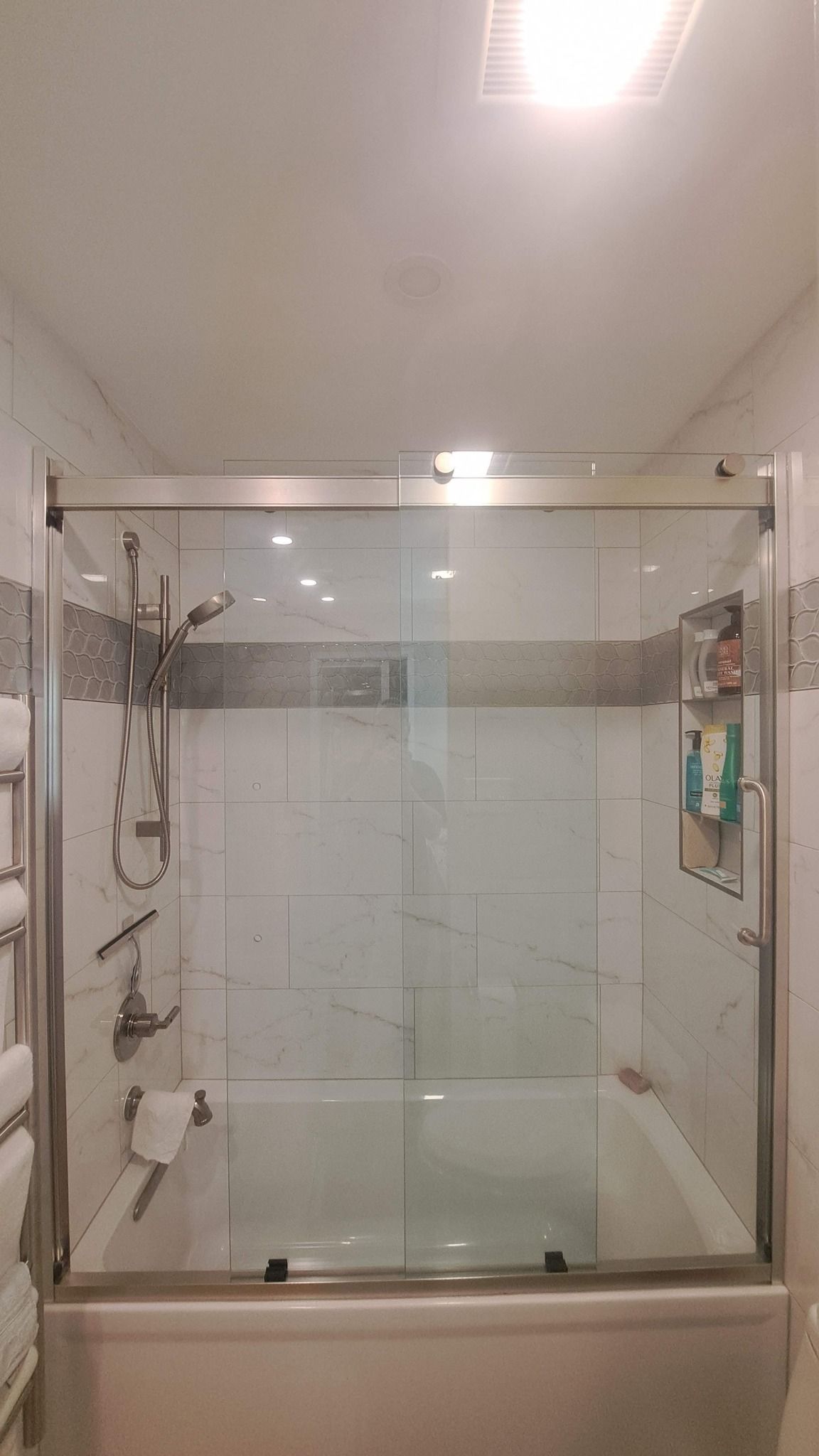 A shower over a bathtub with glass doors, white tile, and a shower head.