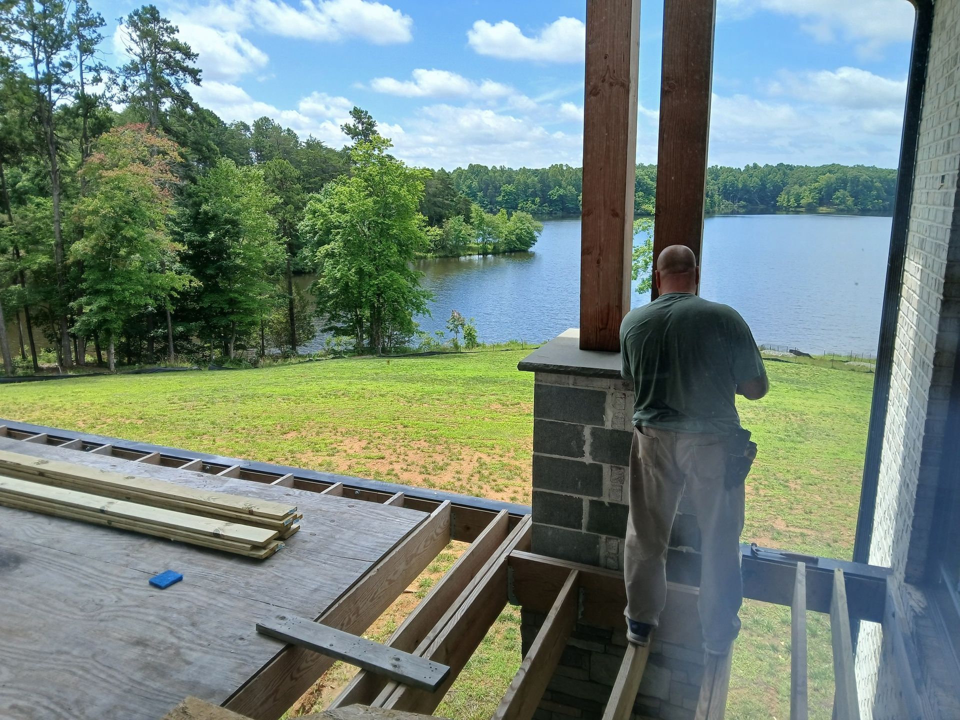 A person works on a partially built deck overlooking a lake and trees on a sunny day.