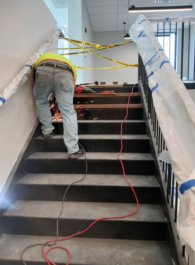 Construction worker cutting material on staircase, yellow safety tape above. Black stairs, red power cord.