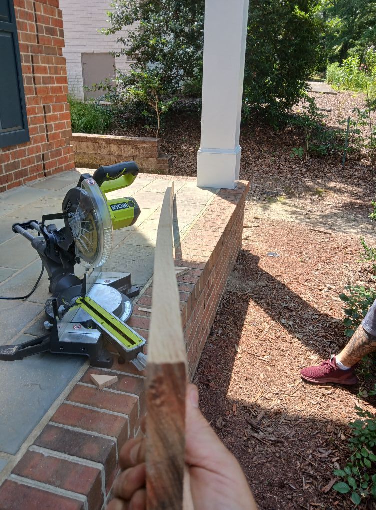 Person holds pointed wooden dowel near a miter saw on a brick porch. Sunny outdoor setting.
