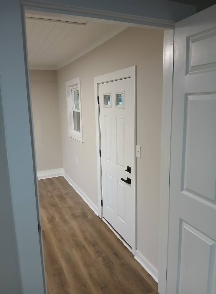 Narrow hallway with white door, window, and wood-look flooring. Light gray walls and ceiling.