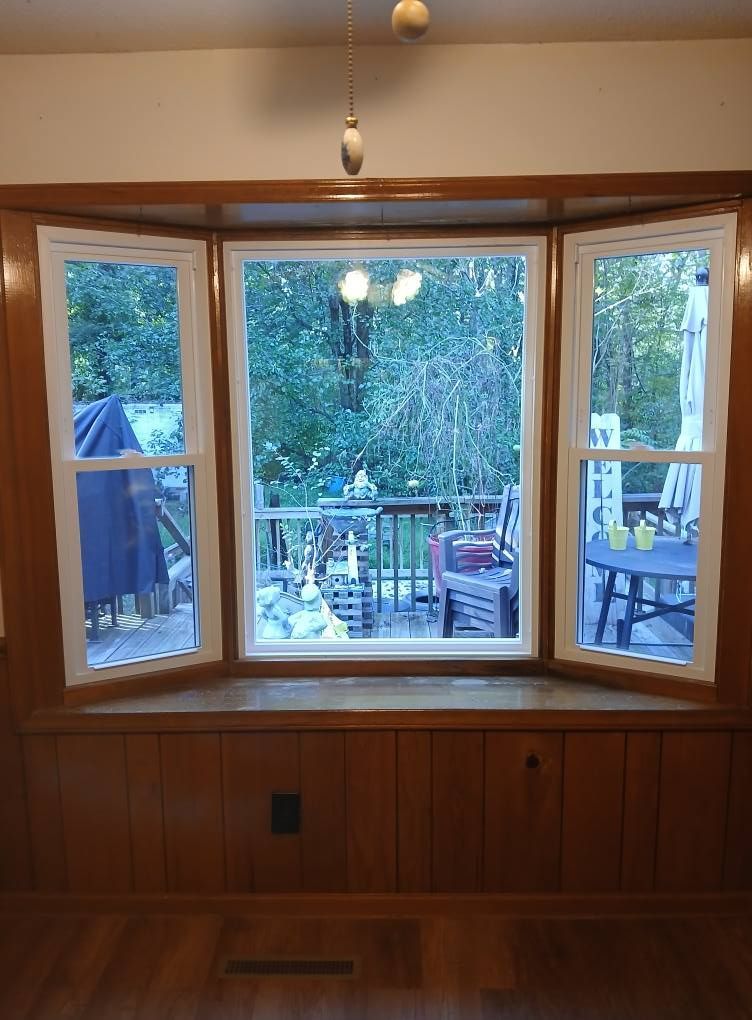 Bay window with white trim, overlooking a deck with furniture, surrounded by trees.