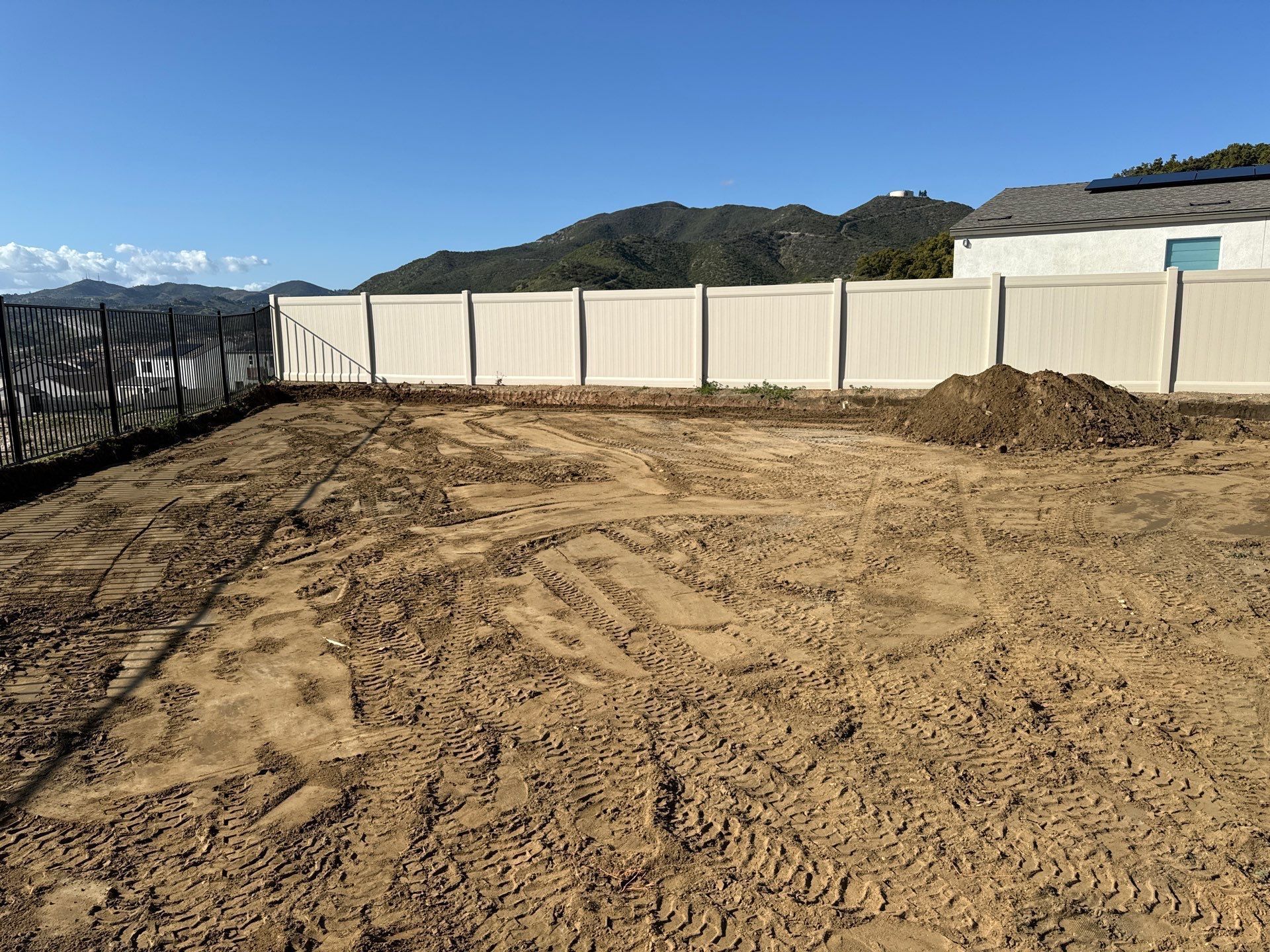 Cleared dirt lot enclosed by white fence with mountains in background.