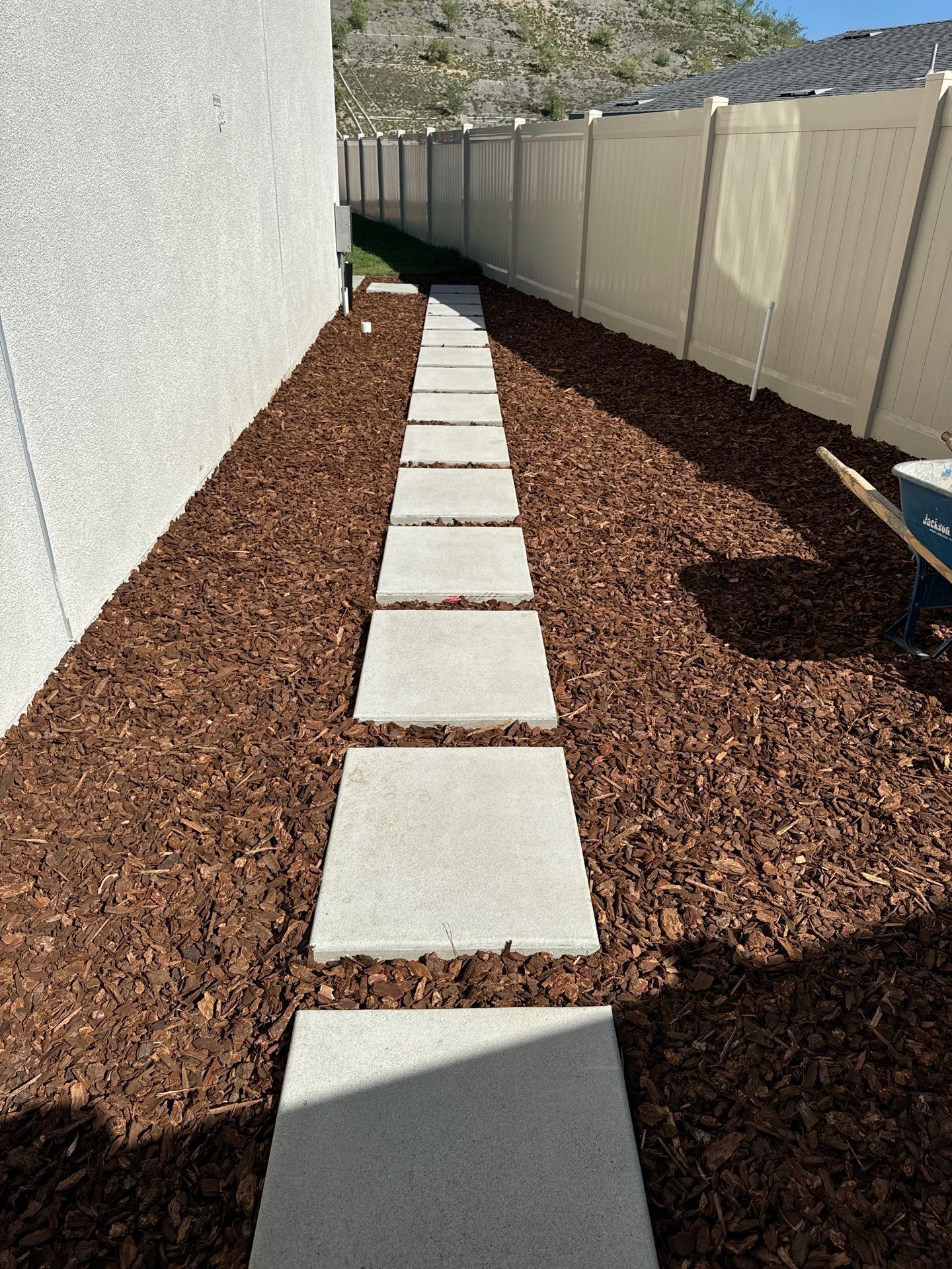 Concrete stepping stones through brown mulch in a narrow yard between a light-colored wall and fence.