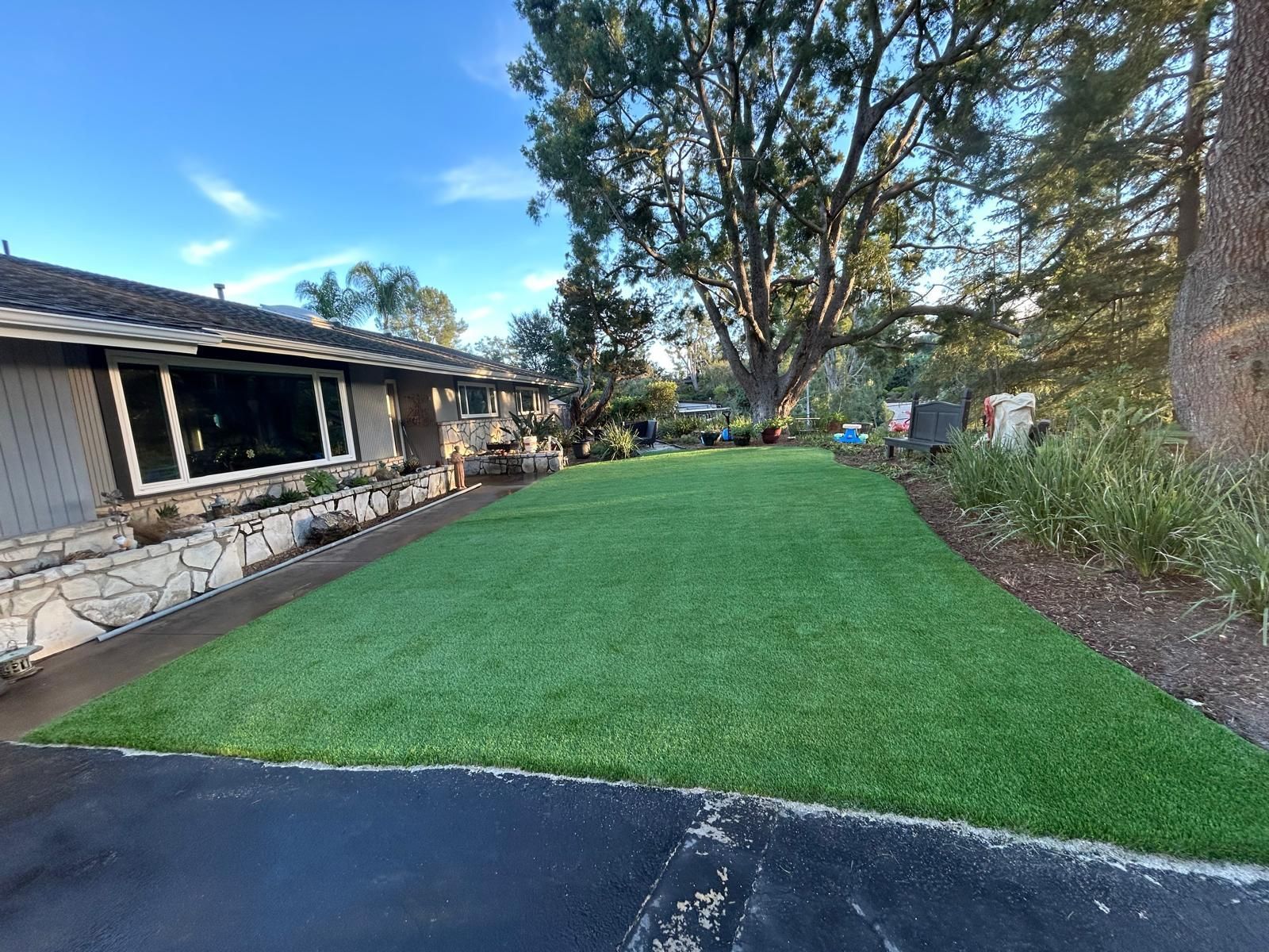 Artificial green turf lawn in front of a house. Dark driveway on the foreground. Trees and blue sky background.