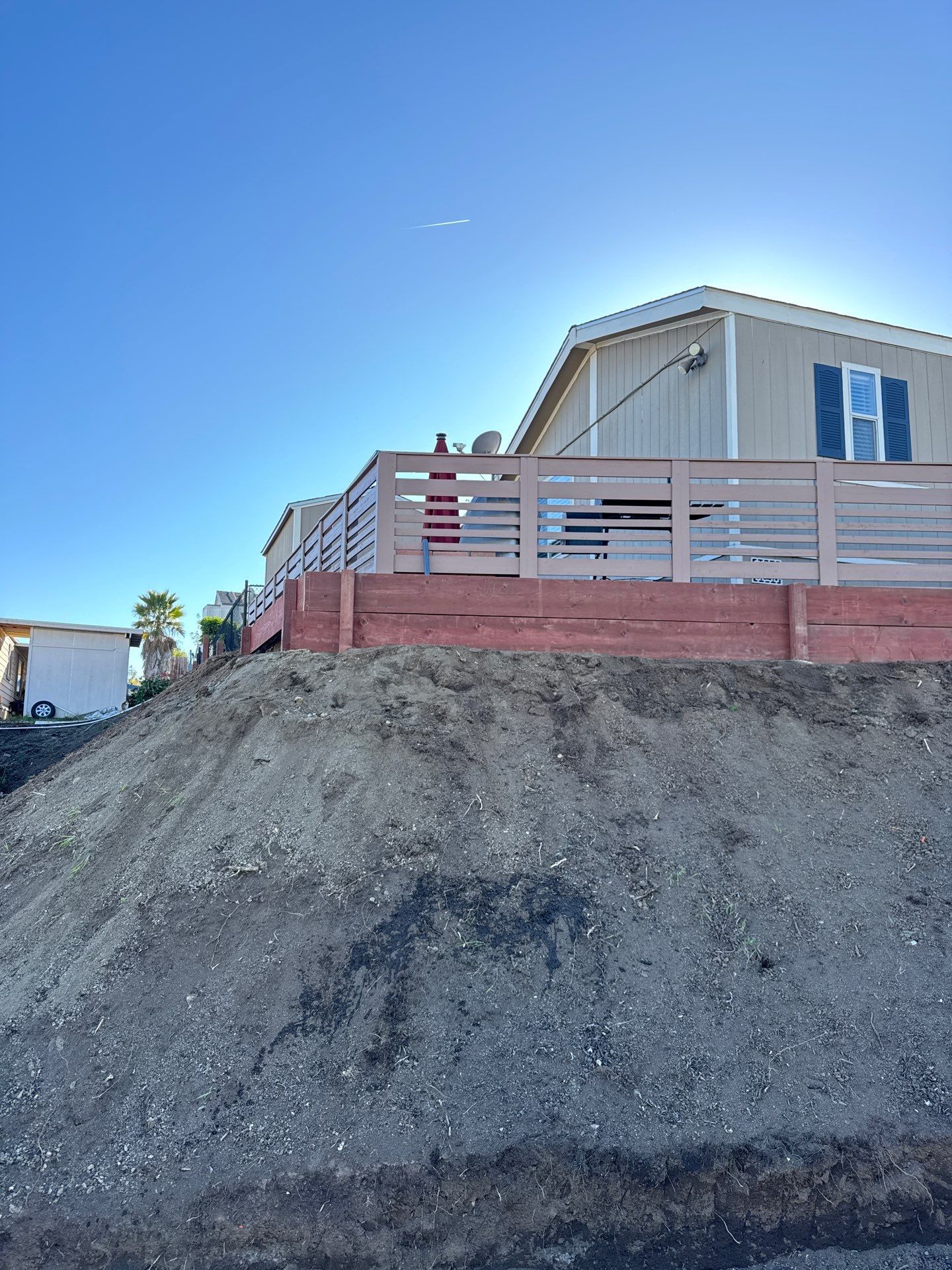 Dirt mound in front of tan mobile home with a wooden deck and blue sky.