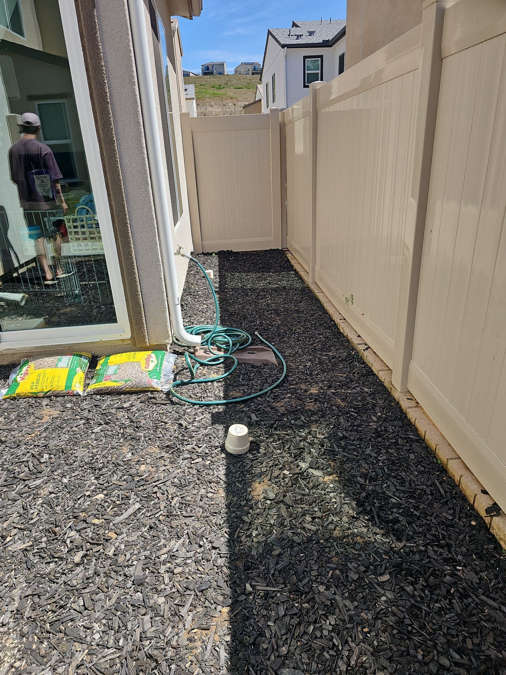 Narrow backyard bed with black mulch, next to a beige fence and house. A hose and person visible.