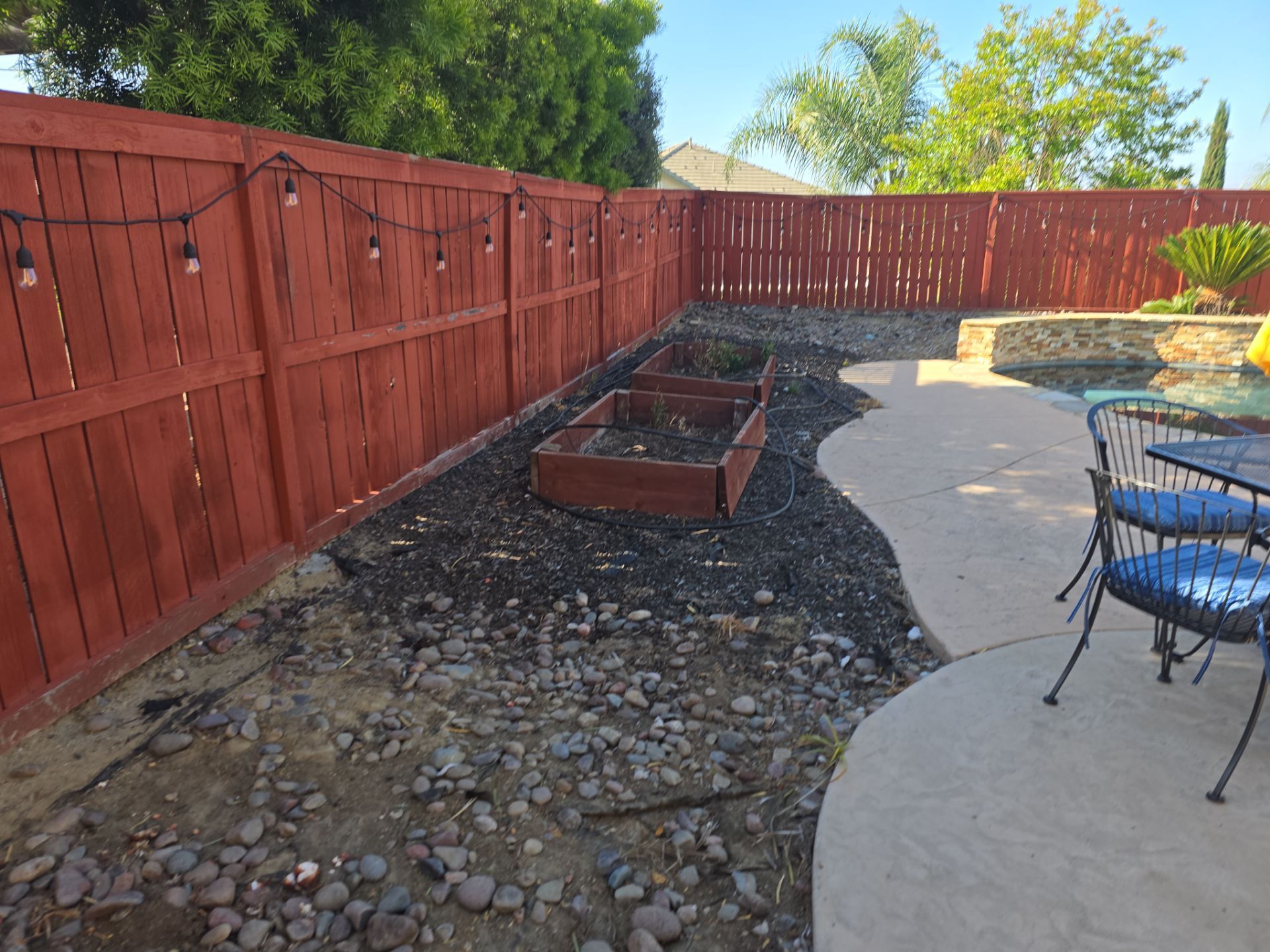 Red-stained wooden fence borders a backyard garden bed filled with dark mulch and rocks, near a pool and patio.