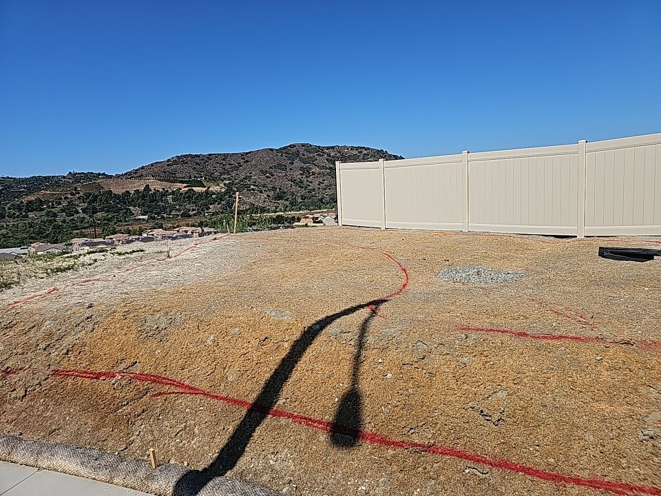 A dirt lot marked with red lines, fenced on the right, with a mountain in the distance under a blue sky.