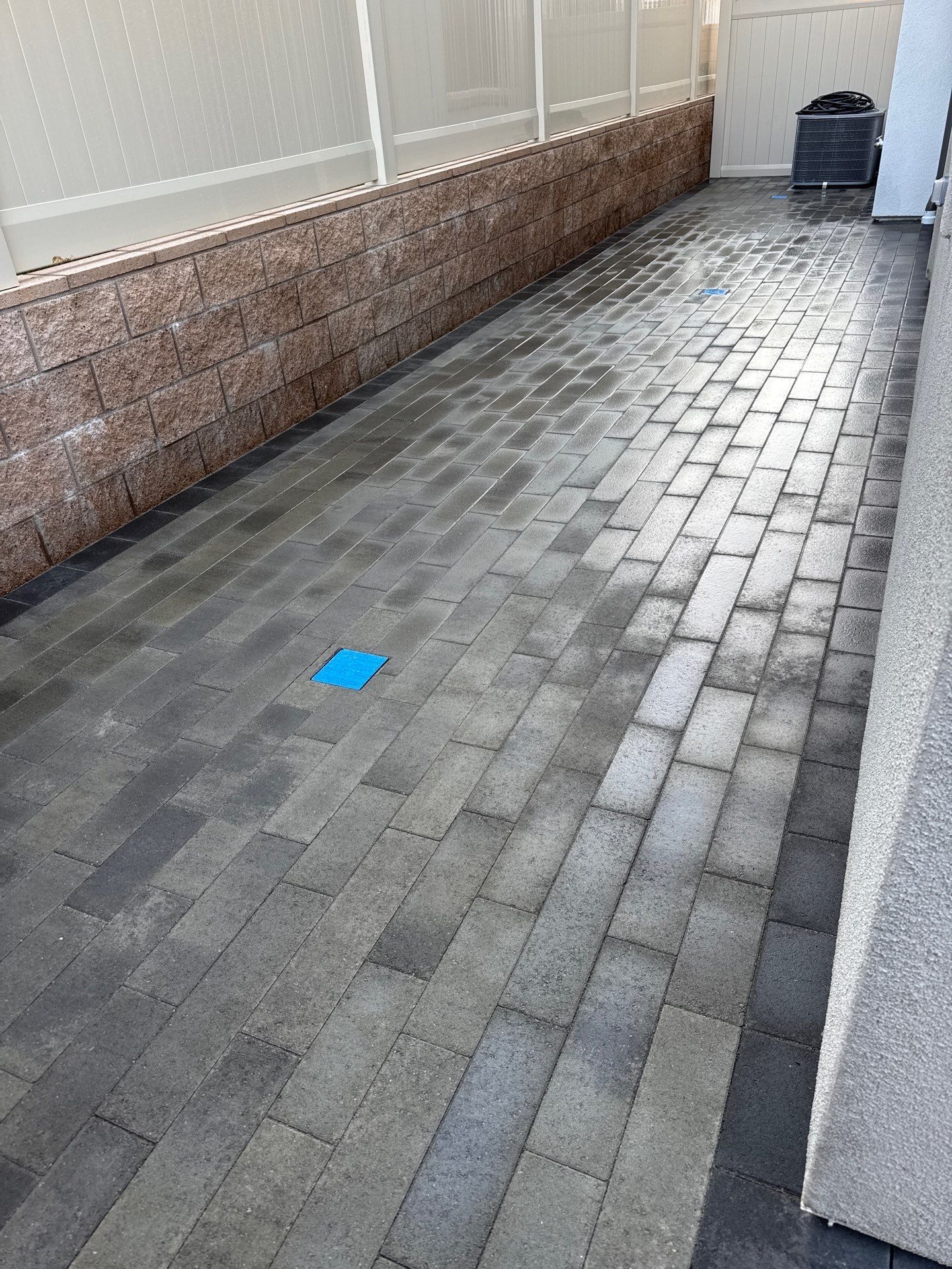 Brick paved walkway next to a brown brick wall and a white fence. Blue square on the ground.