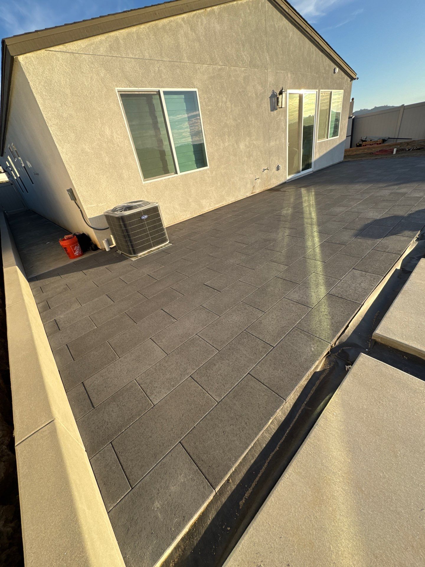 Backyard patio with dark gray pavers, light tan retaining wall, and beige house with AC unit.