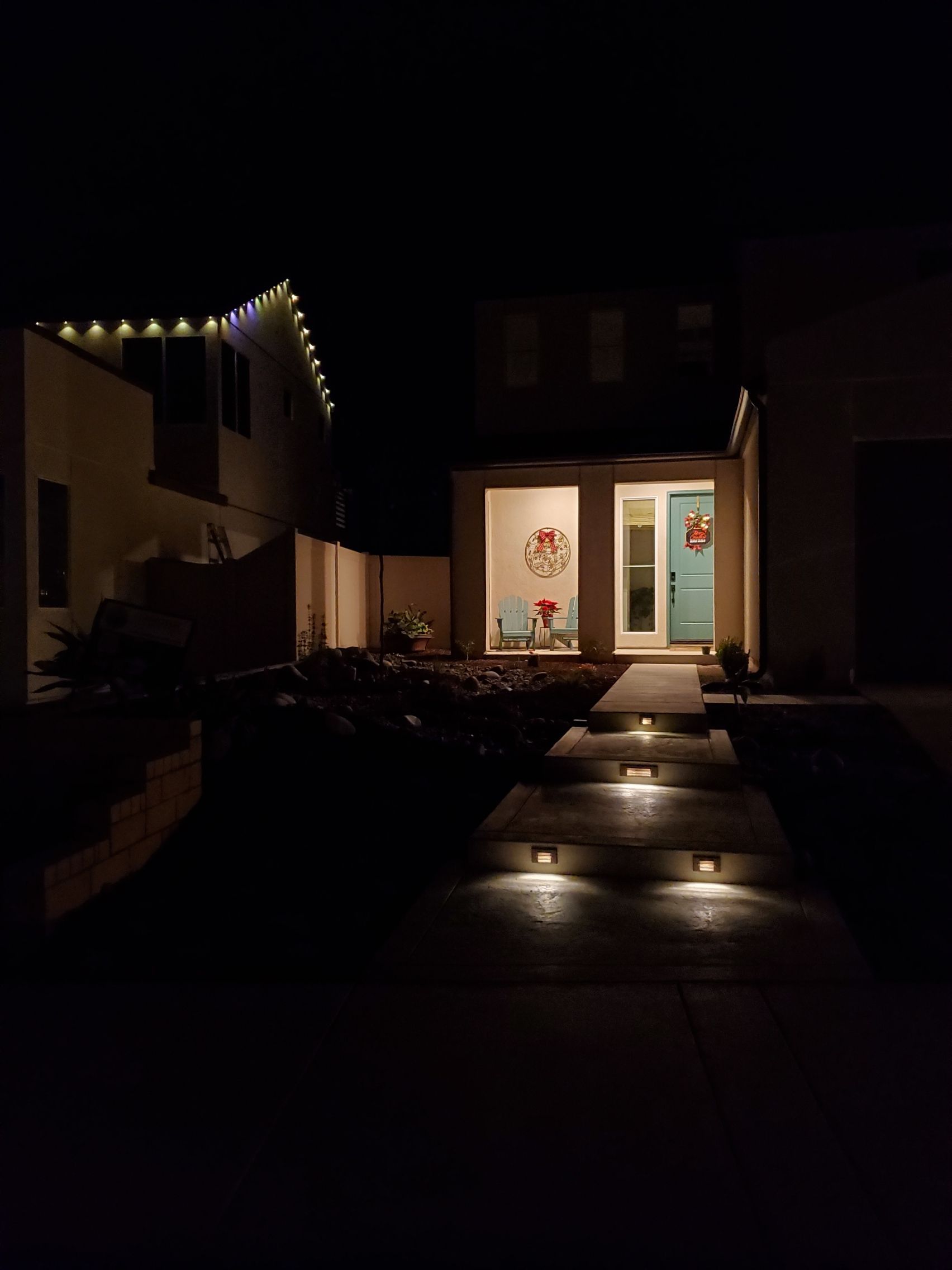 Night shot of a house with exterior lights on. Lit doorway and pathway leading to the front door, with Christmas lights.