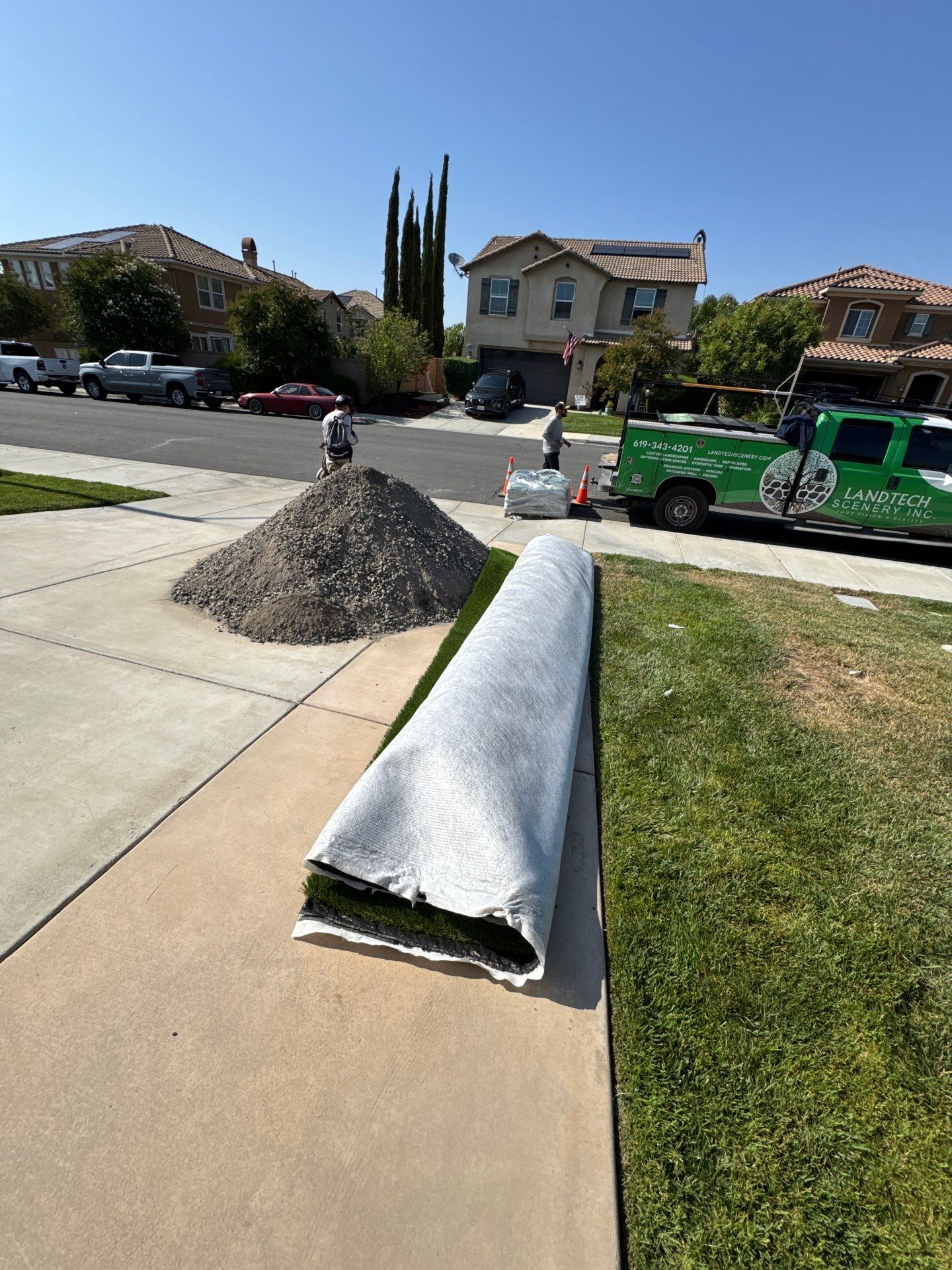 Laying artificial turf on a residential lawn. Workers, gravel pile, rolled turf, green truck parked in street.