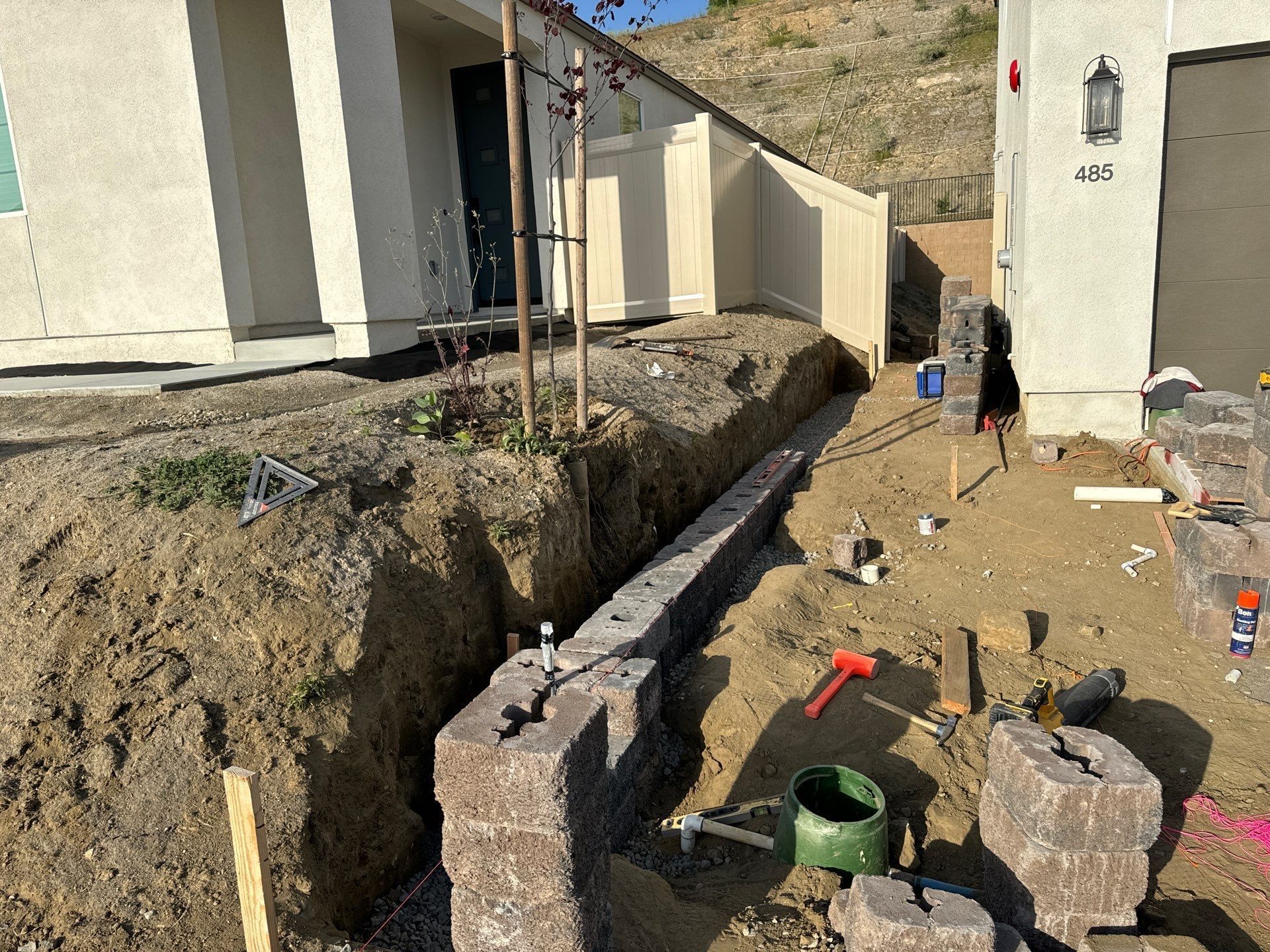 Construction site with retaining wall being built next to a house. Dirt, blocks, tools, and a fence are visible.