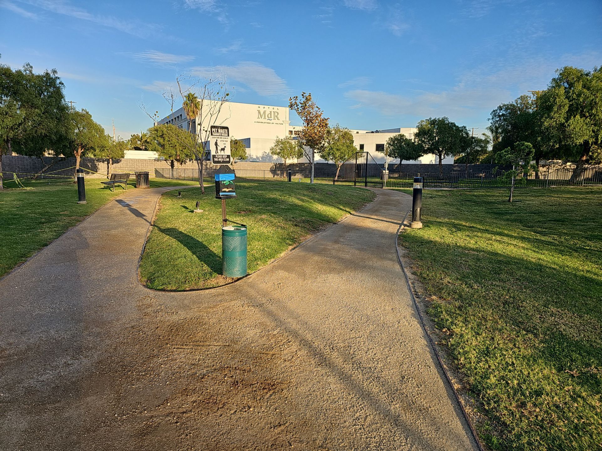 Park with paved paths, grass, trees, and a building in the background. A trash can and sign are in the center.