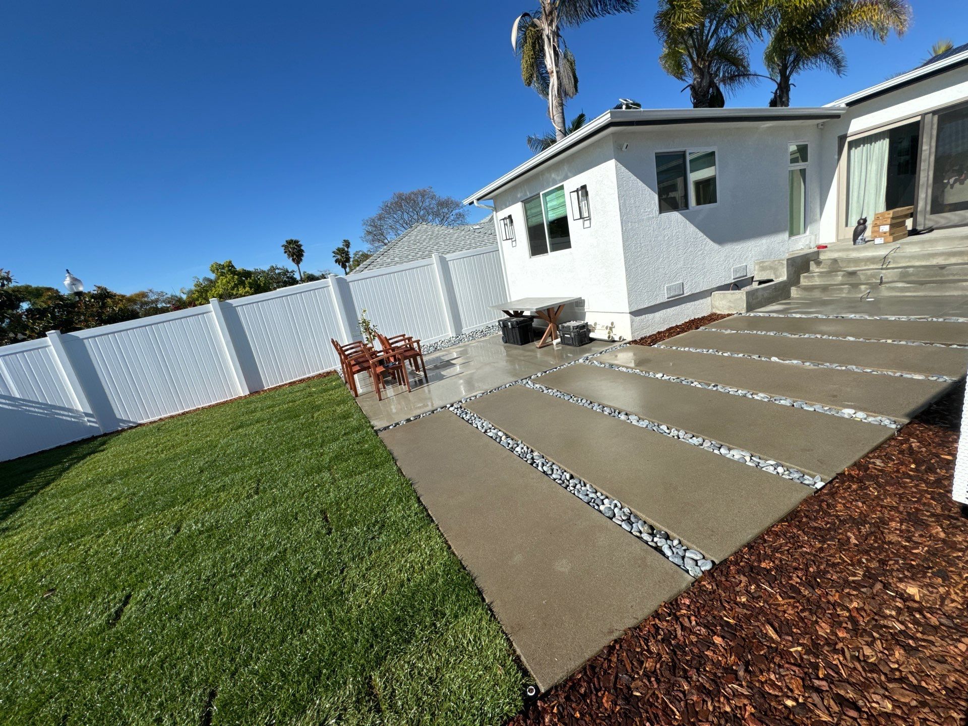 Backyard with concrete pavers, gravel, white fence, and green grass against a white house under a blue sky.