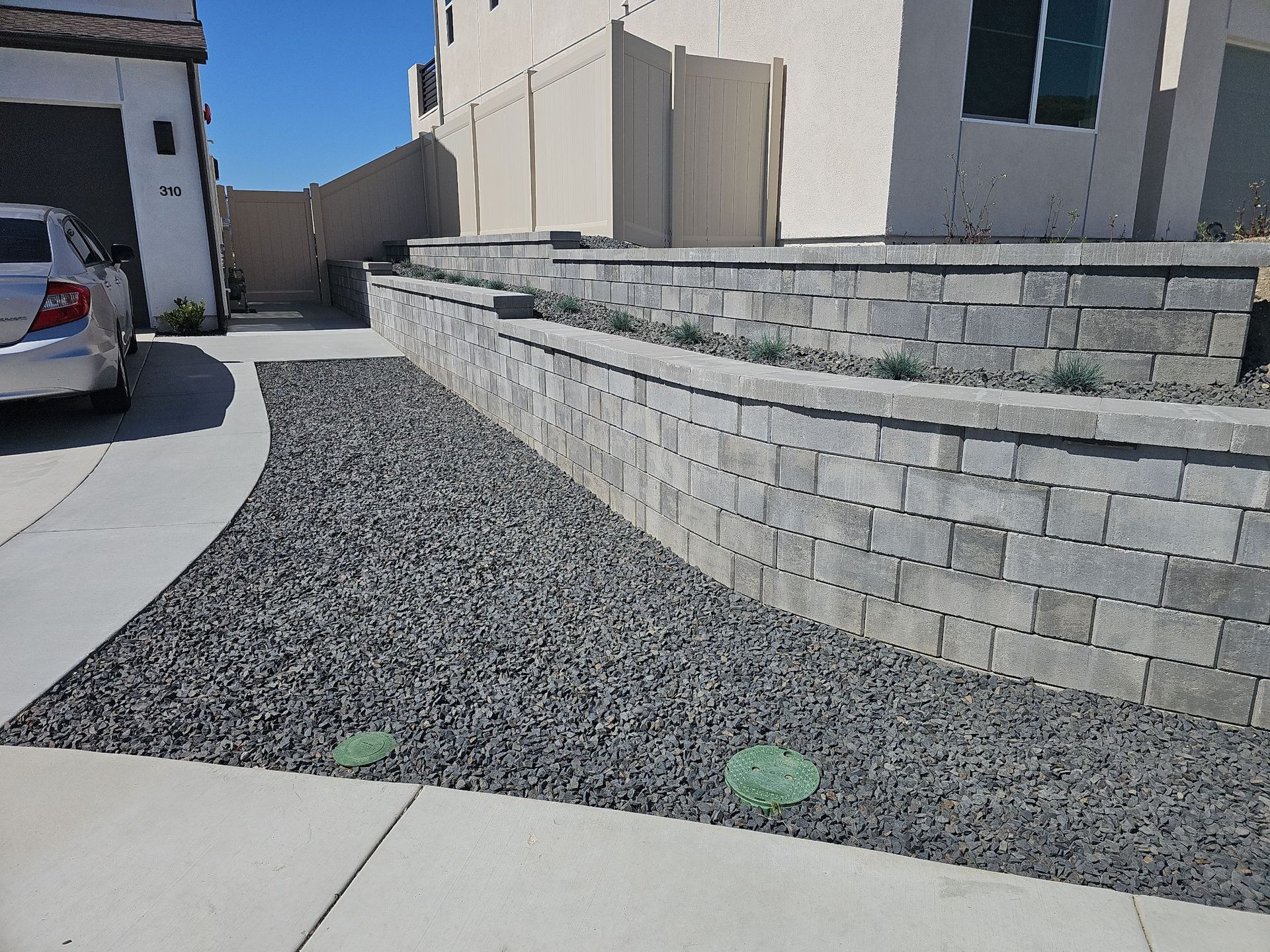 Grey brick retaining walls in front of a house, with gravel and greenery. A car is parked nearby.