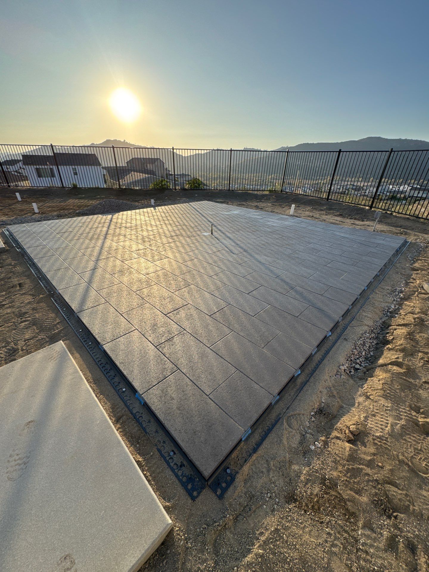 Square black flooring installed on a dirt plot, with a fence and sunlit sky in the background.
