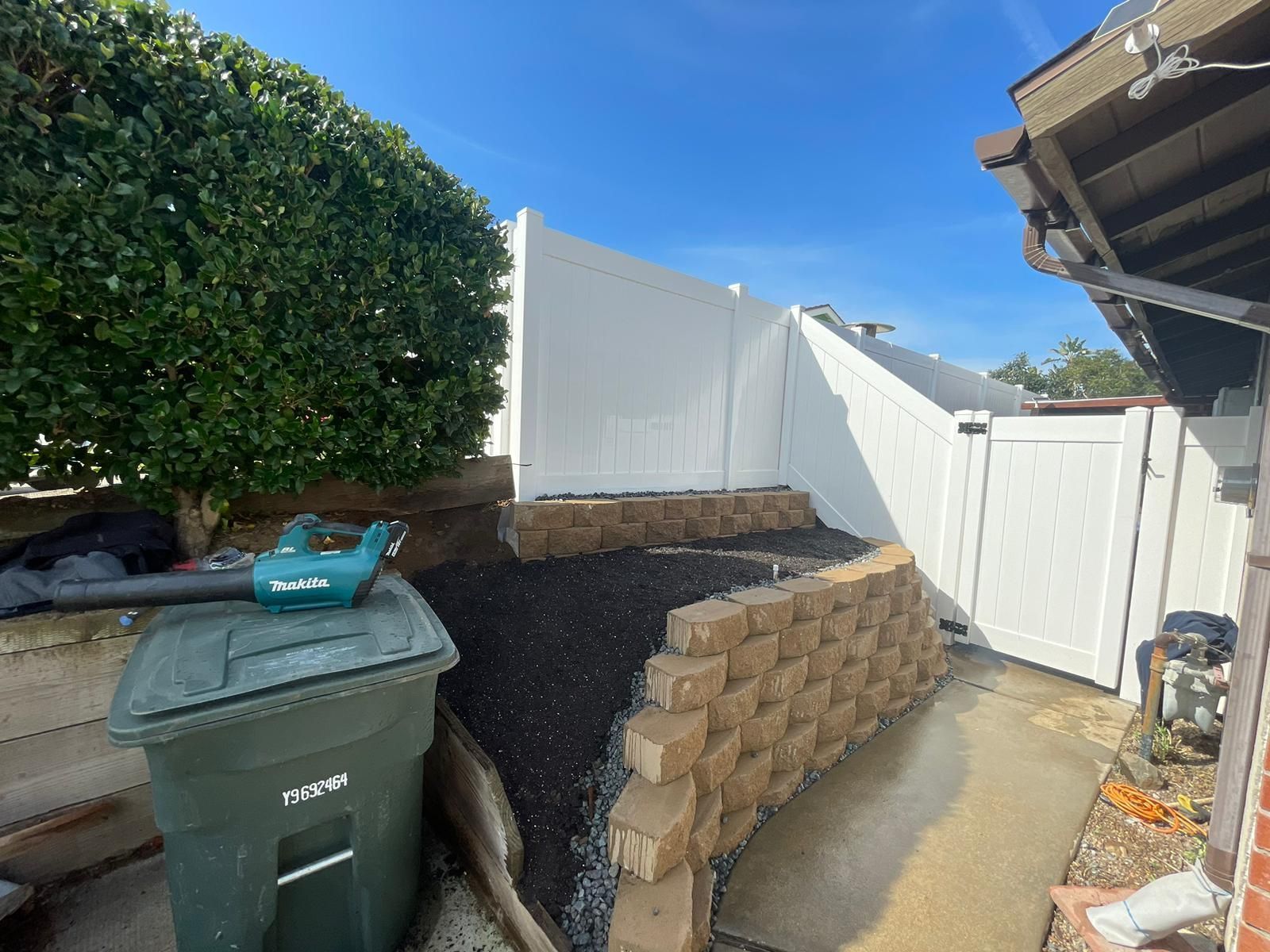 White fence with stacked retaining wall, green trash bin, and blower in a backyard under a blue sky.